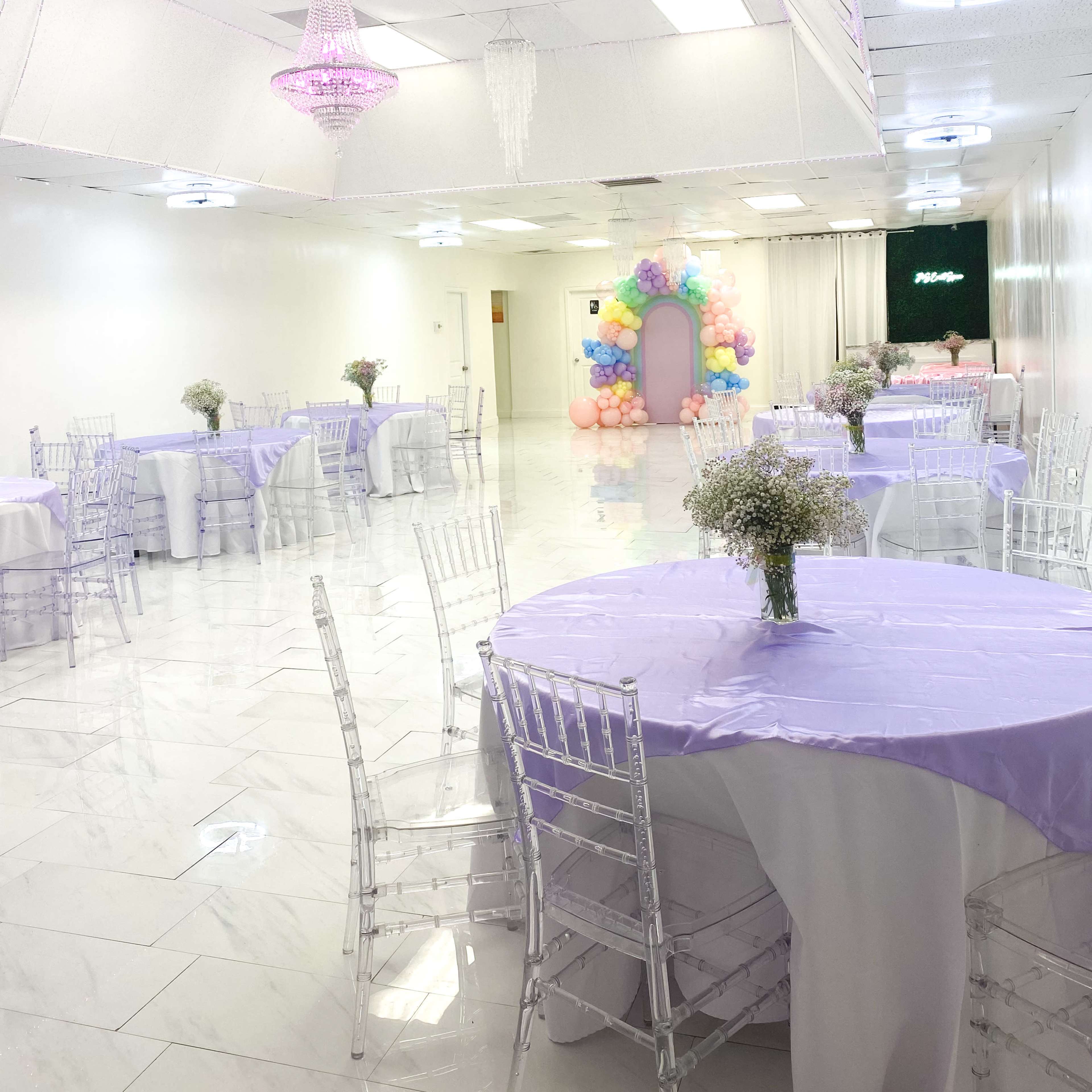 A spacious event hall decorated with round tables covered in light purple tablecloths, clear chiavari chairs, and a colorful balloon arch in the background.