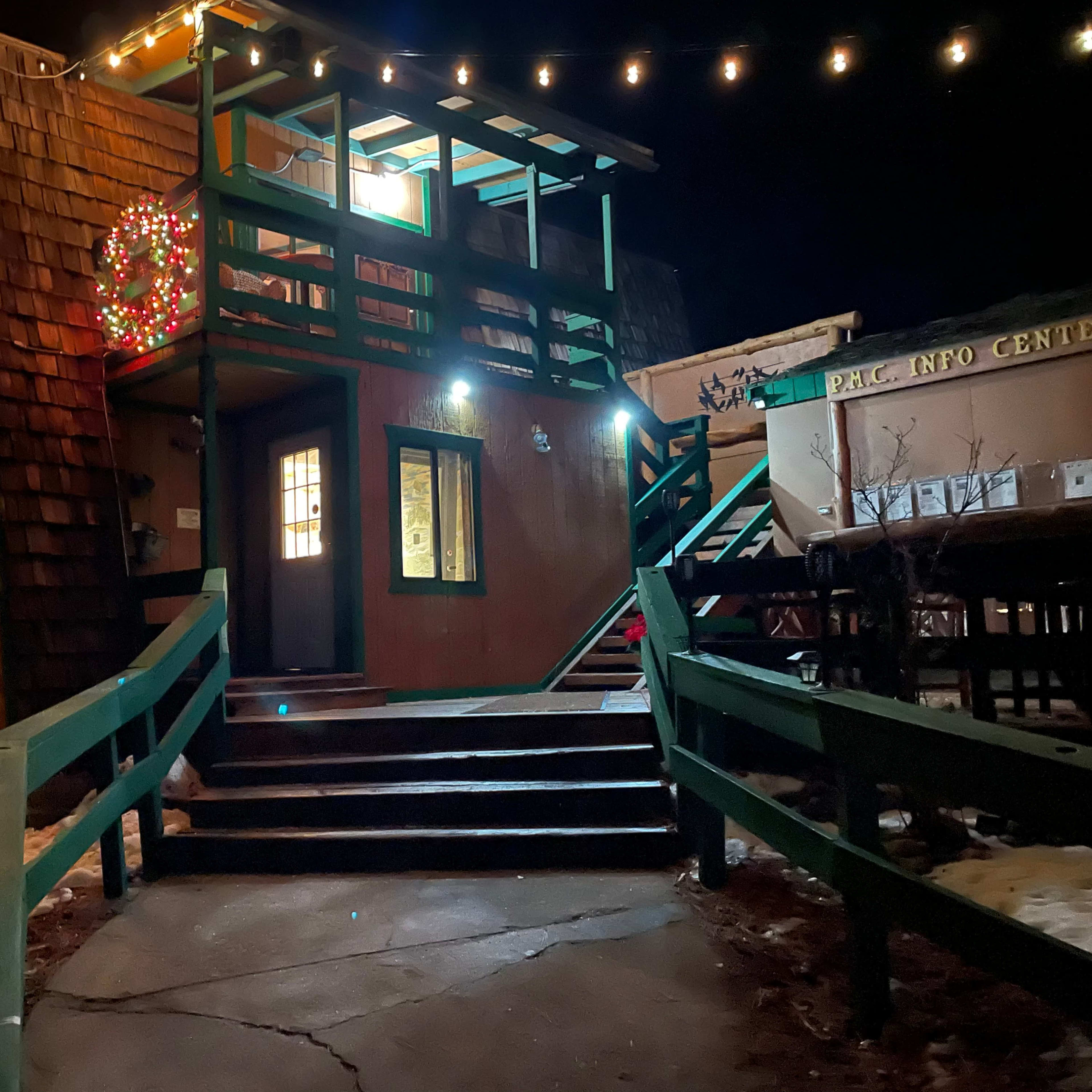 A two-story building with green railings and Christmas lights is illuminated at night, surrounded by patches of snow.