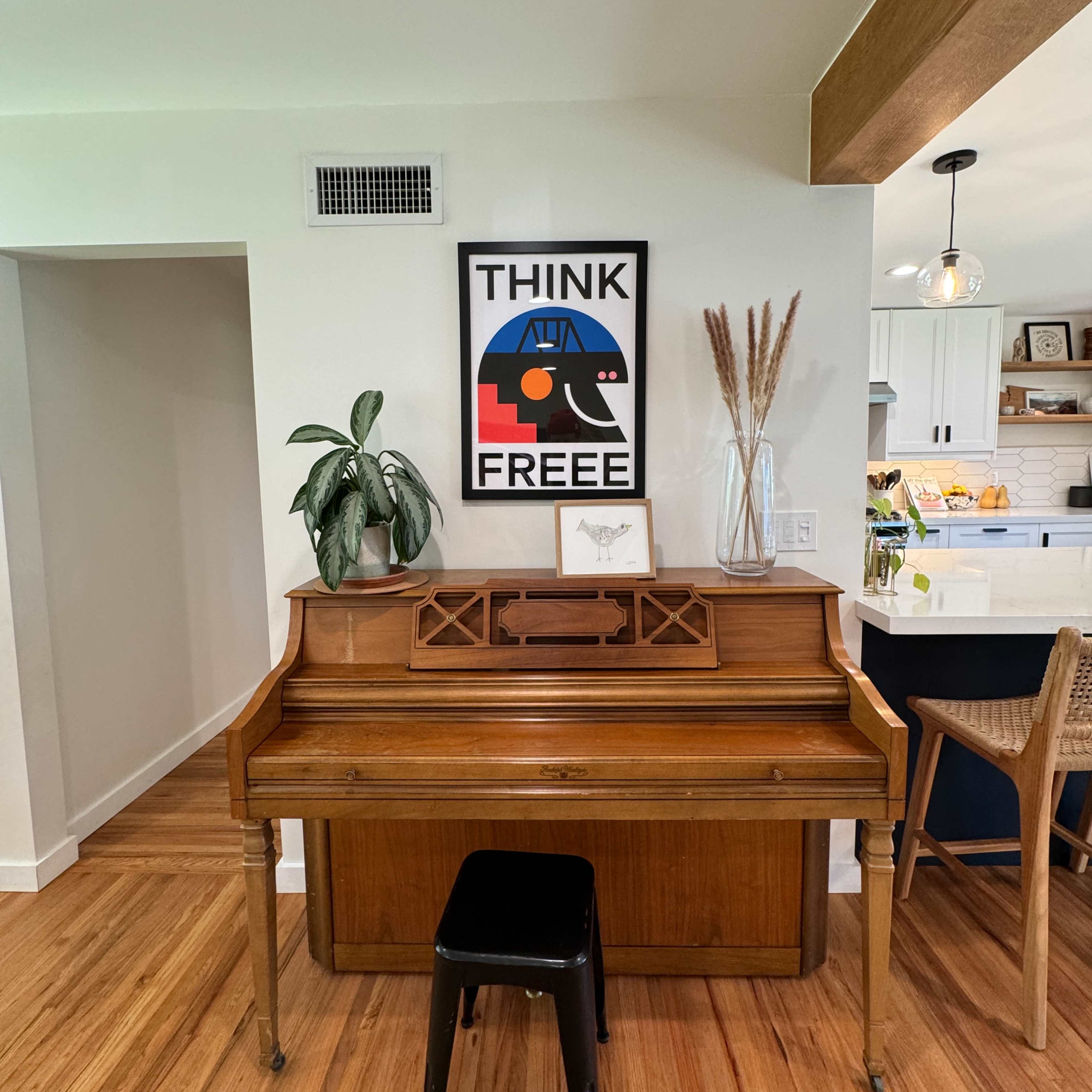 A vintage wooden piano is positioned against a wall featuring a framed poster that reads "THINK FREE," with decorative plants and a glass vase nearby.