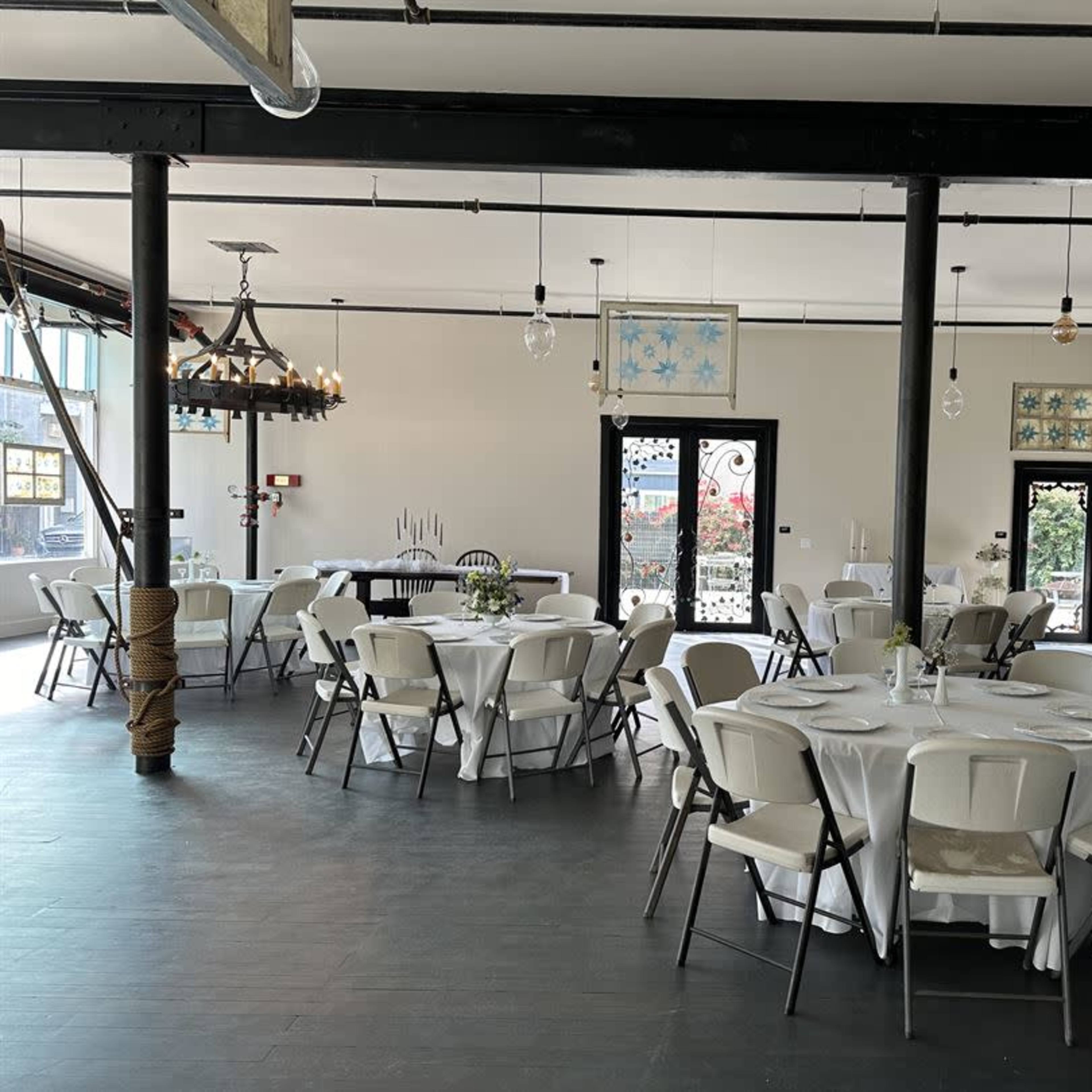 A spacious event room with several round tables set with white tablecloths and chairs, along with decorative elements and large windows.