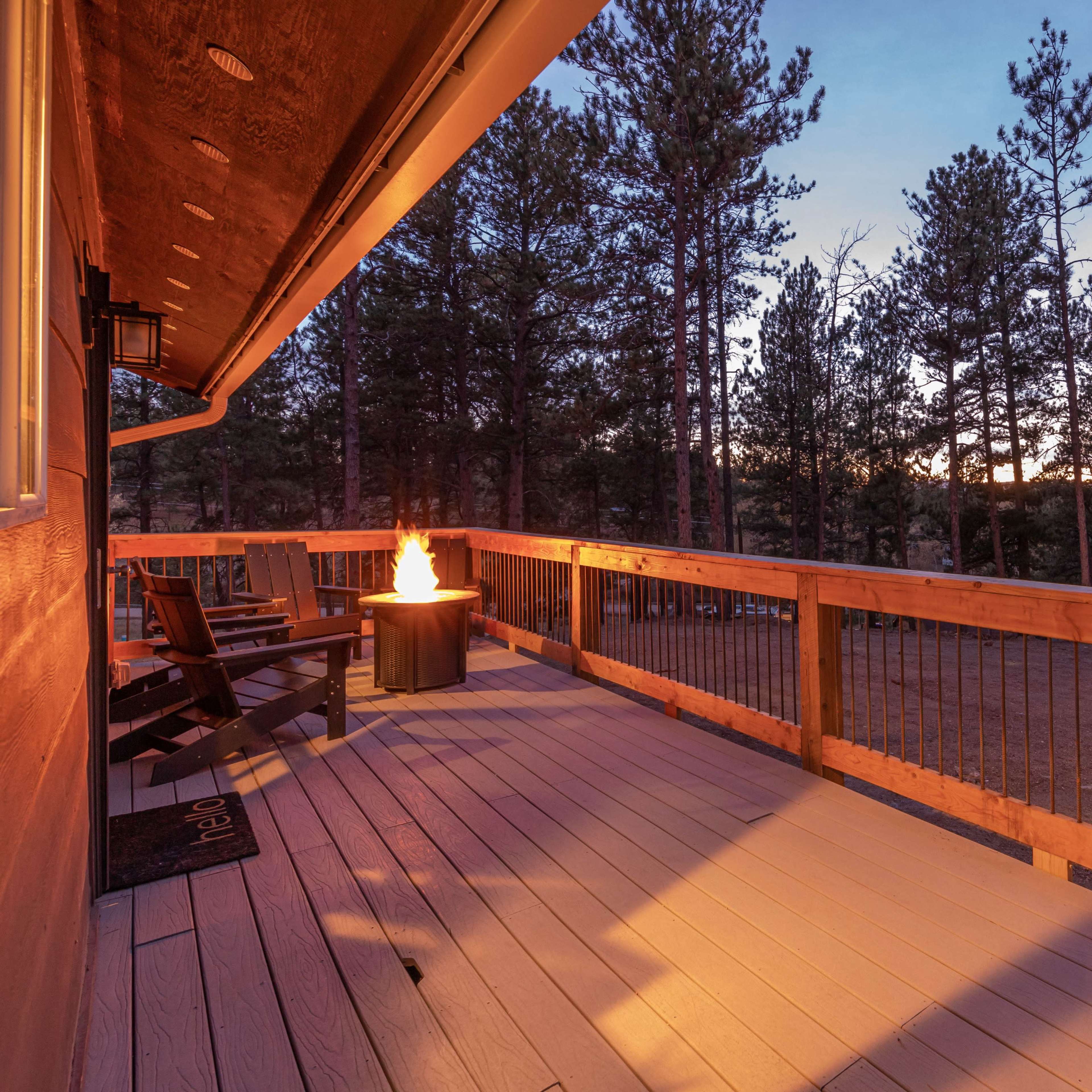 A wooden deck with two chairs and a small fire pit, surrounded by tall trees against a twilight sky.