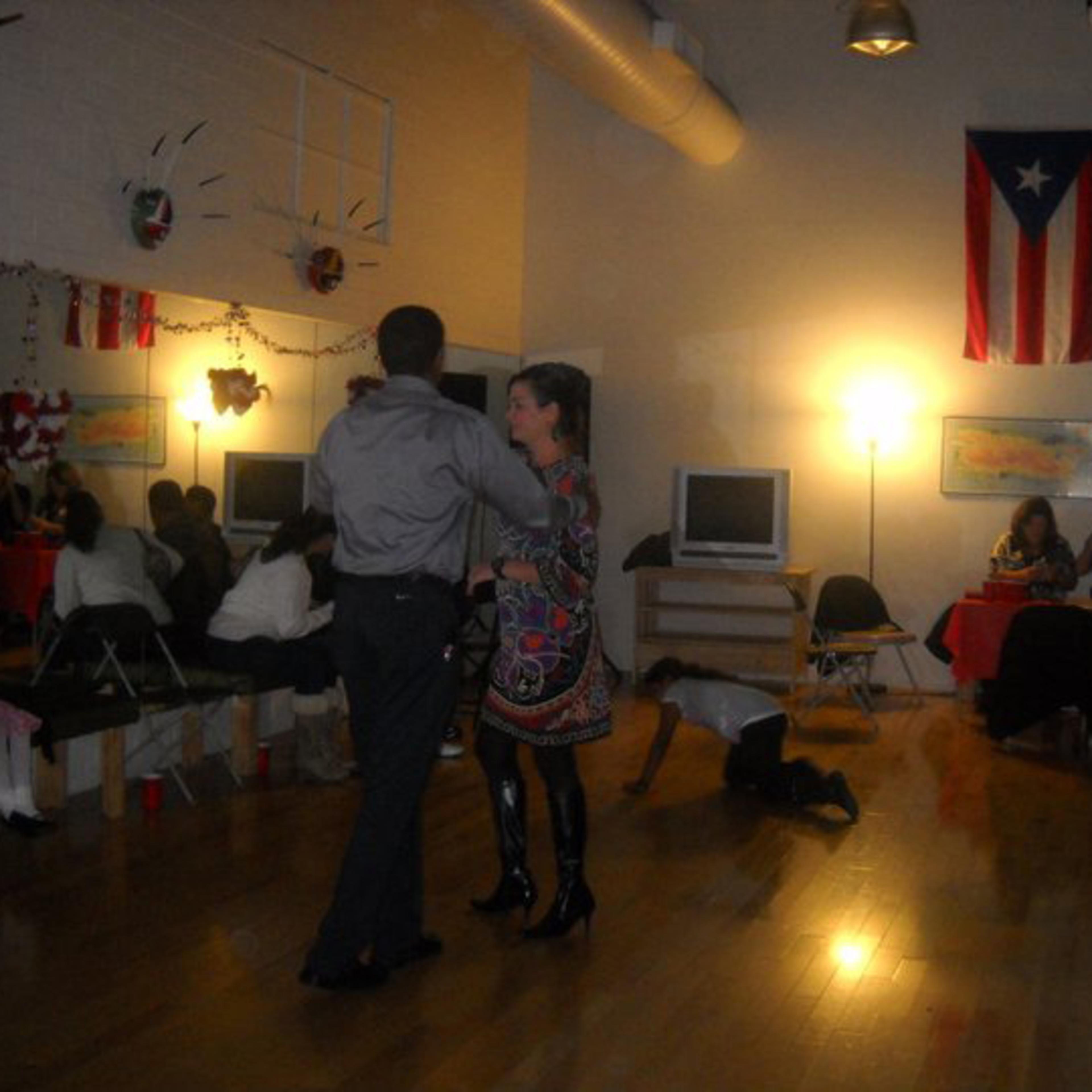 A couple dances in a community center decorated with Puerto Rican flags, while others sit at tables and children play in the background.