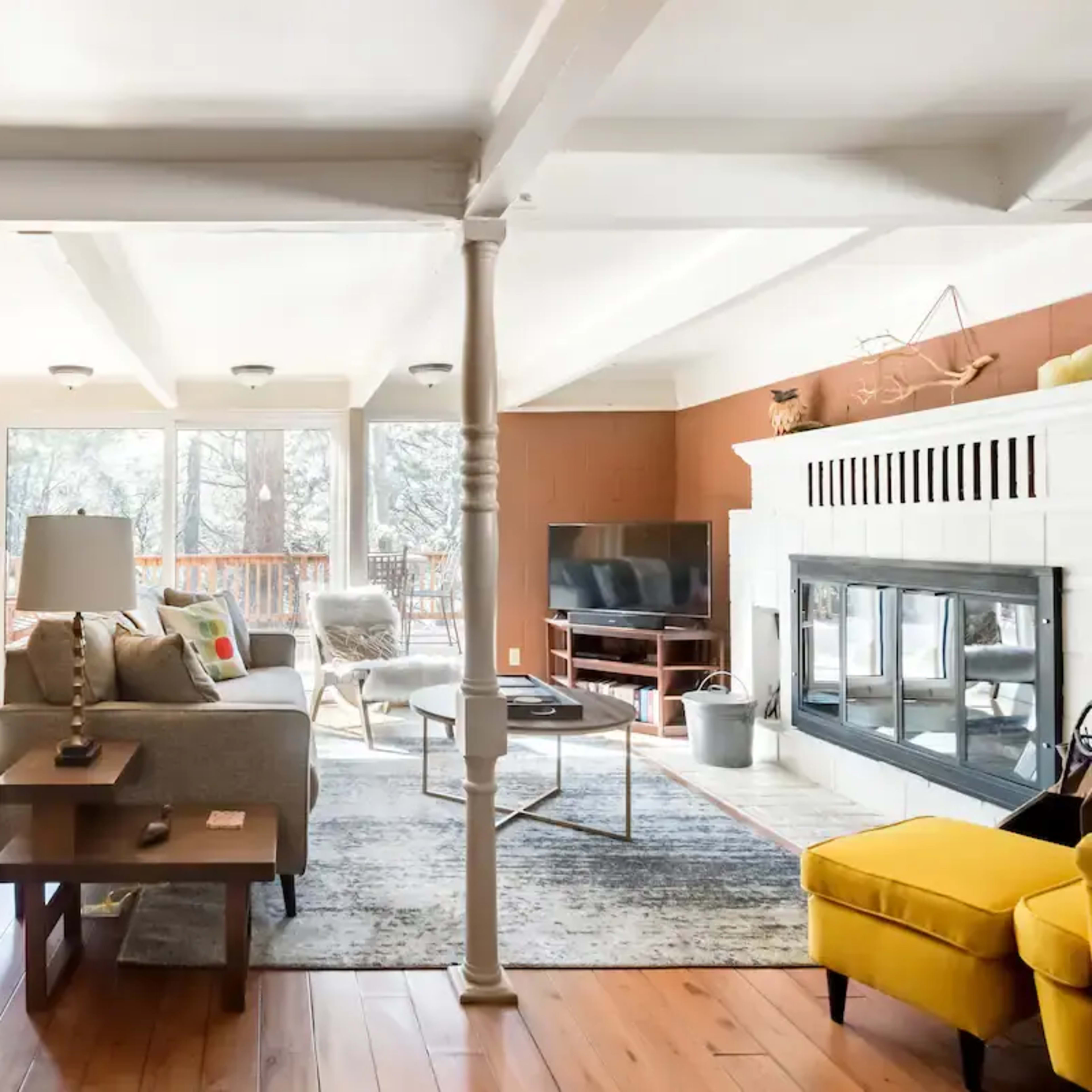 The living room features a neutral-colored sofa, wooden flooring, large windows, and a fireplace, creating an open and airy atmosphere.