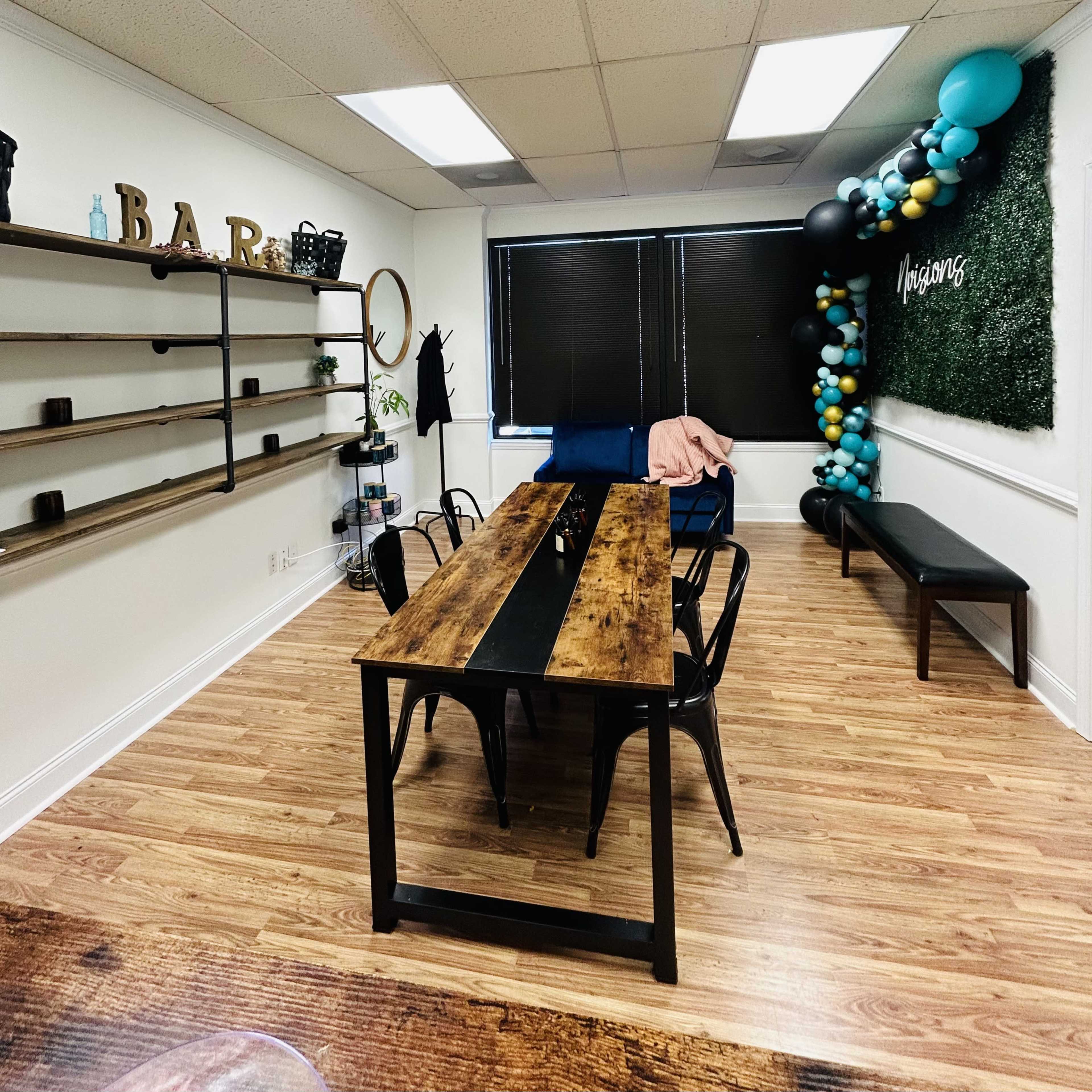 A spacious room with wooden flooring, featuring a long dining table surrounded by black chairs, shelves on one wall, and decorative balloon arrangements in the corner.