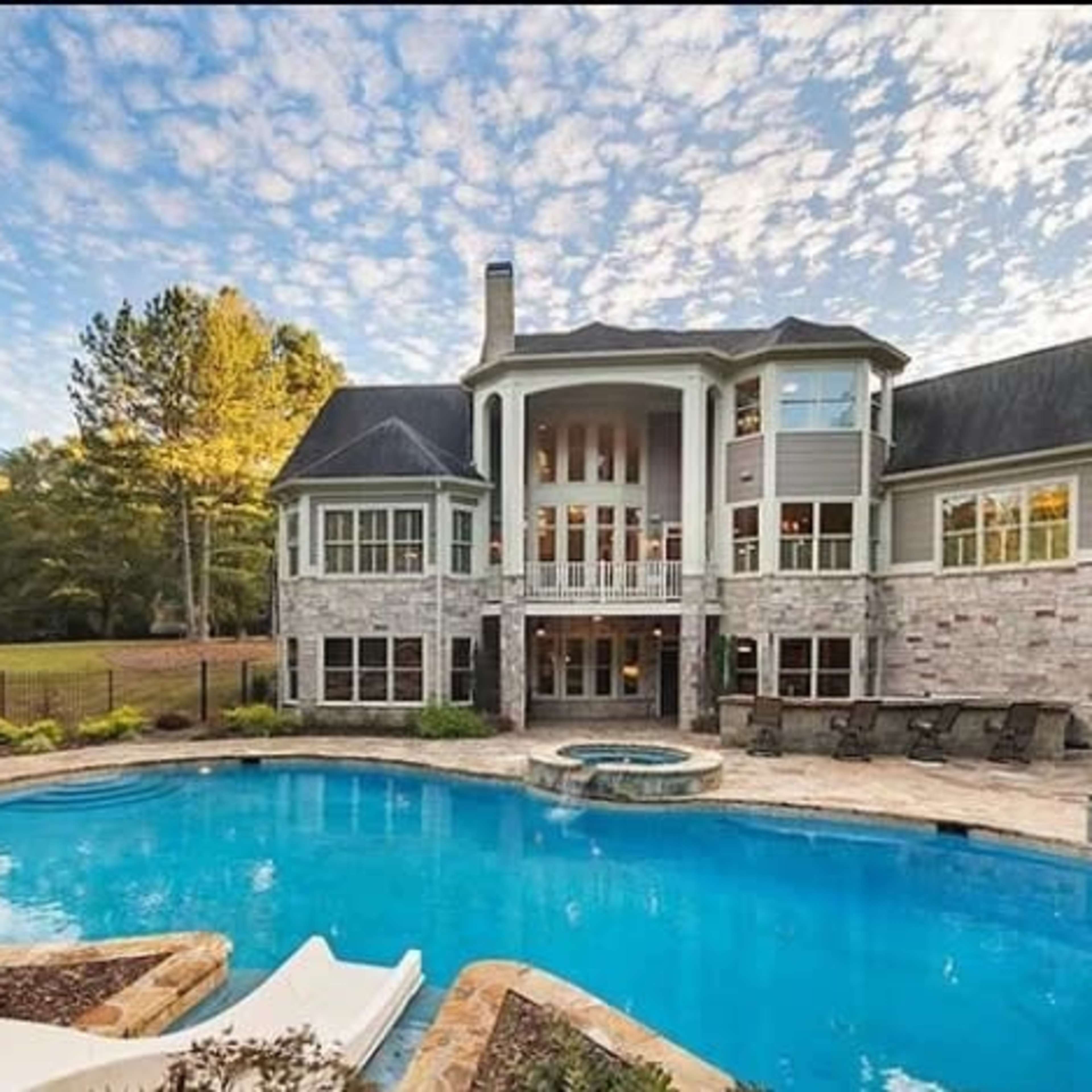 A large stone and wood house with multiple balconies overlooks a blue swimming pool surrounded by landscaped gardens.