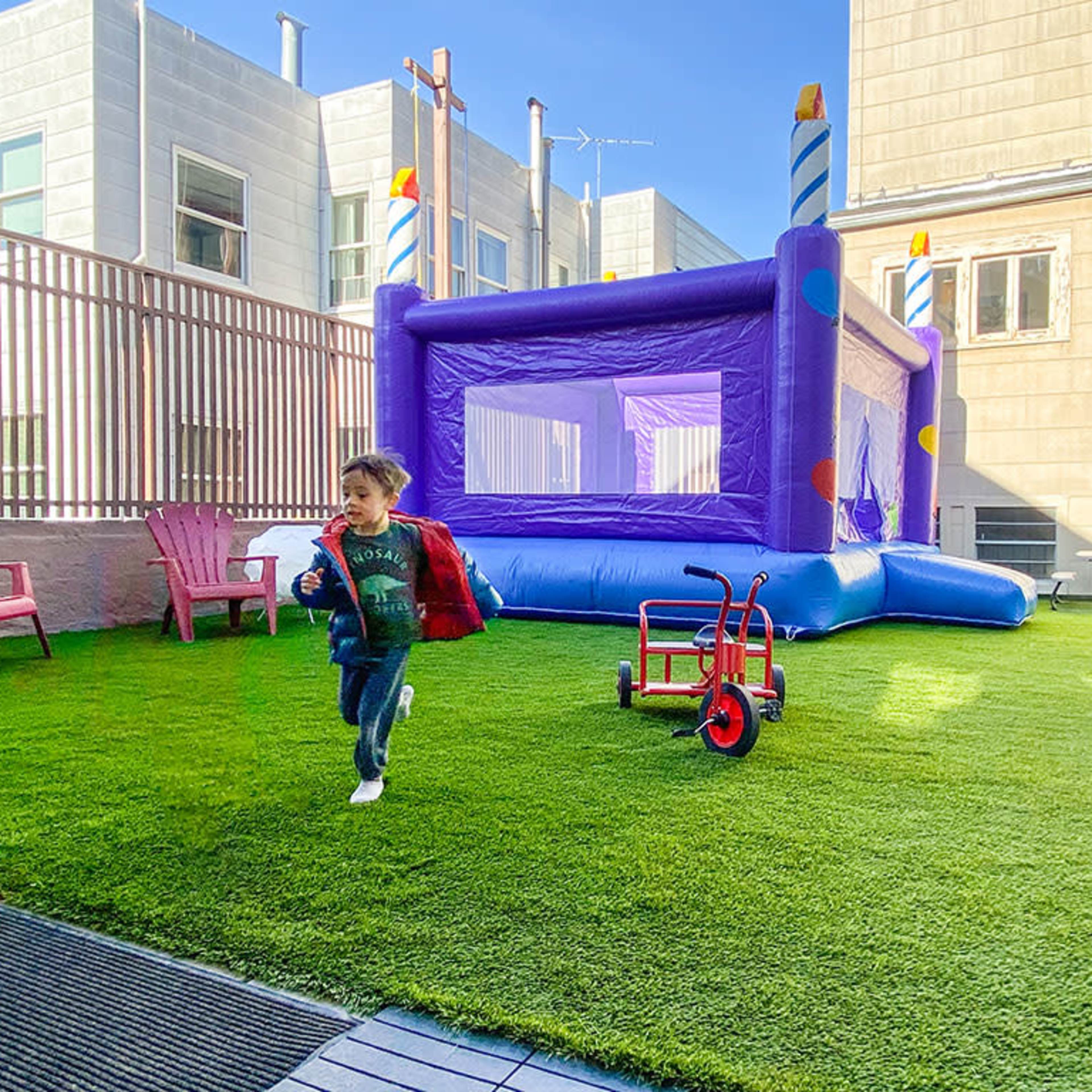A child runs across a grassy area with a purple bounce house and a red tricycle nearby.