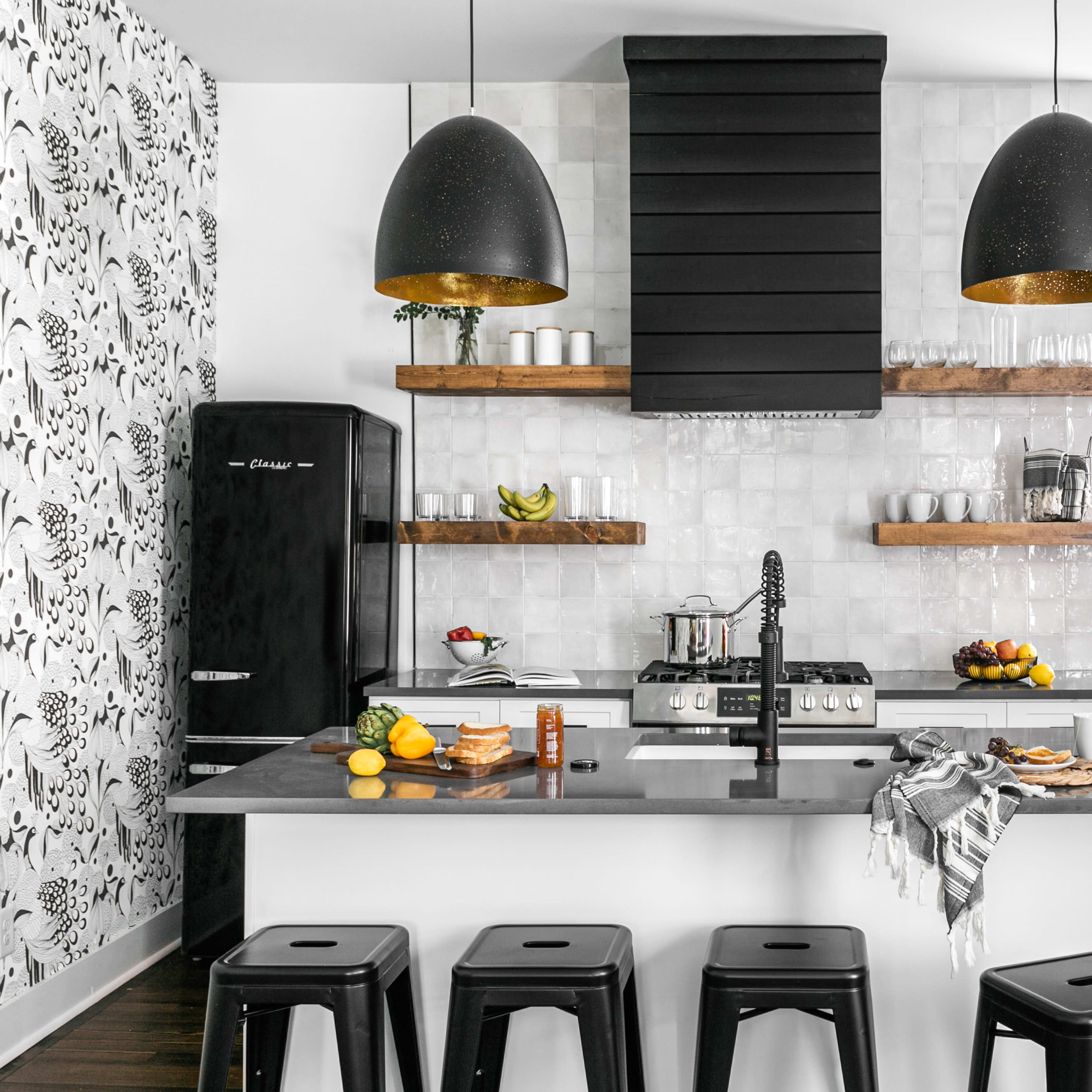 A modern kitchen with black cabinetry, a central island with bar stools, and open shelving displaying kitchenware and fruits.