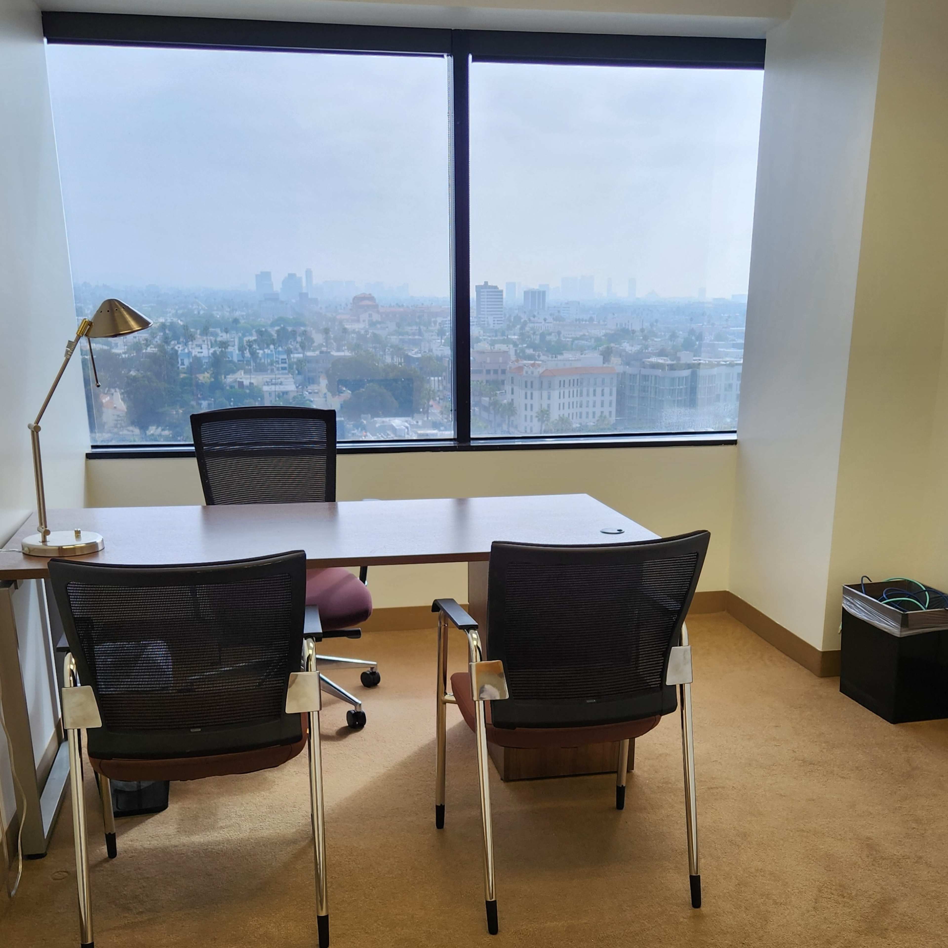 An office space with a large window offering a view of a city skyline, two chairs positioned at a table, and a lamp on the desk.