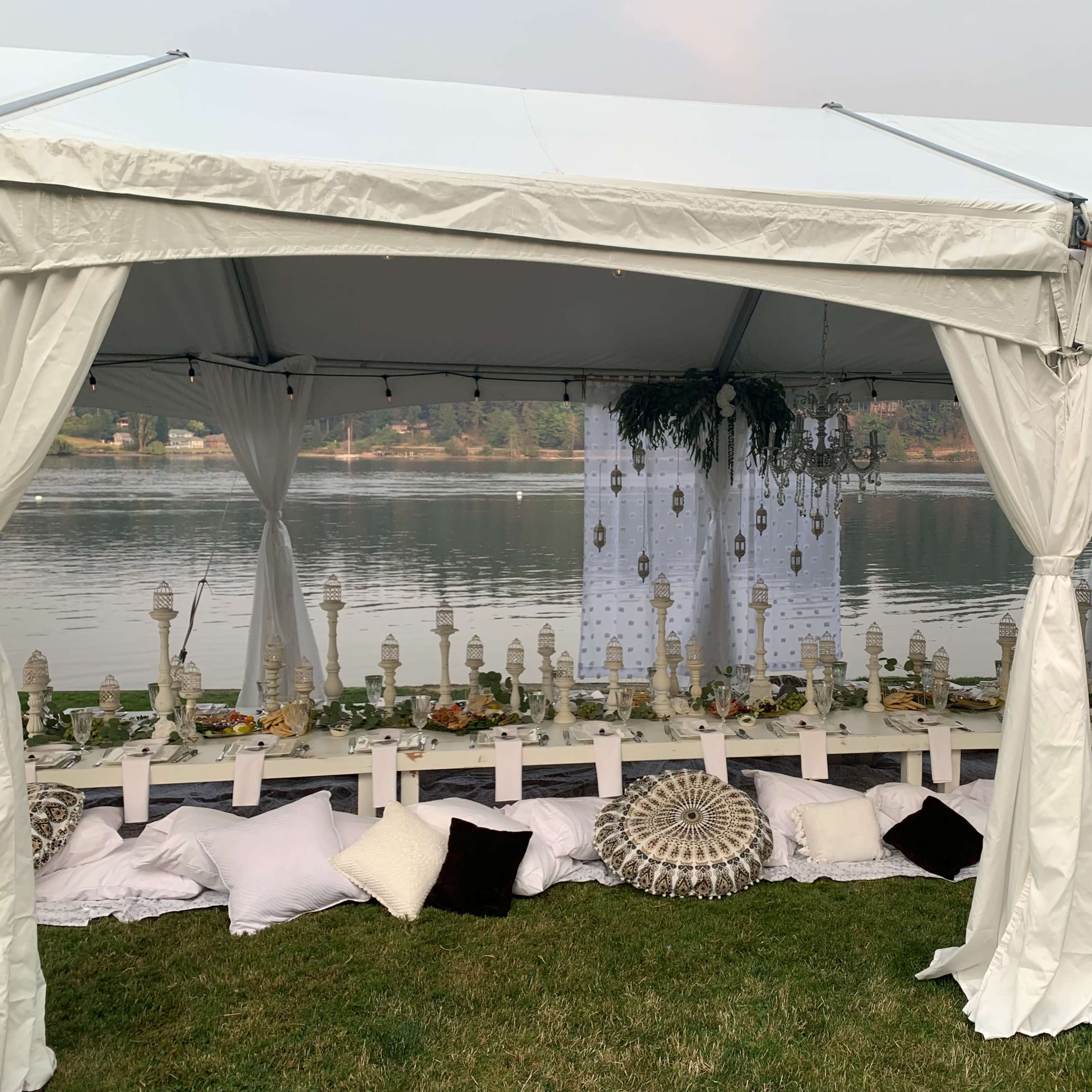 A large white tent is set up by a calm body of water, featuring a long dining table decorated with candles and flowers, surrounded by cushions on the grass.