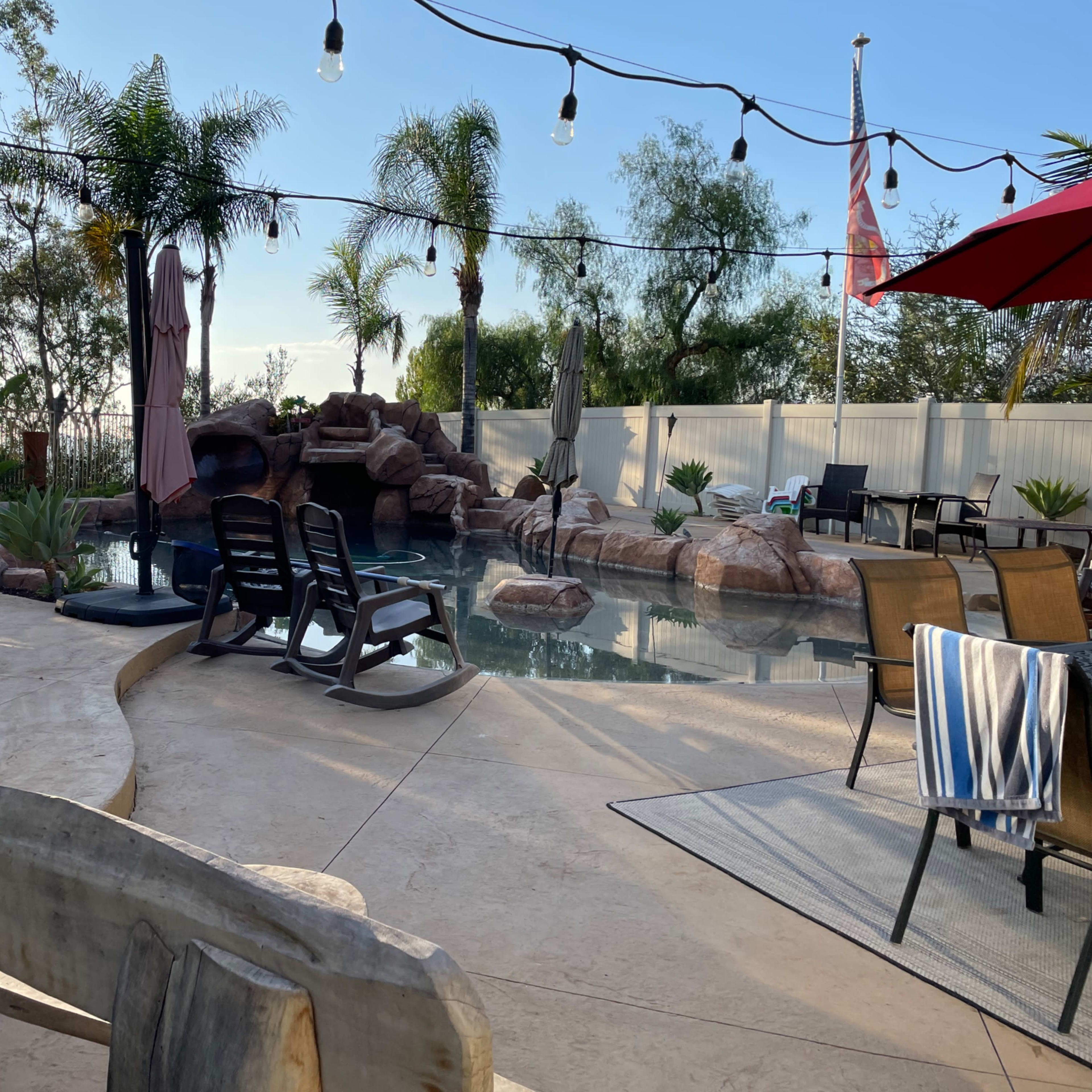 A backyard pool area featuring lounge chairs, a rock waterfall, palm trees, and string lights overhead.
