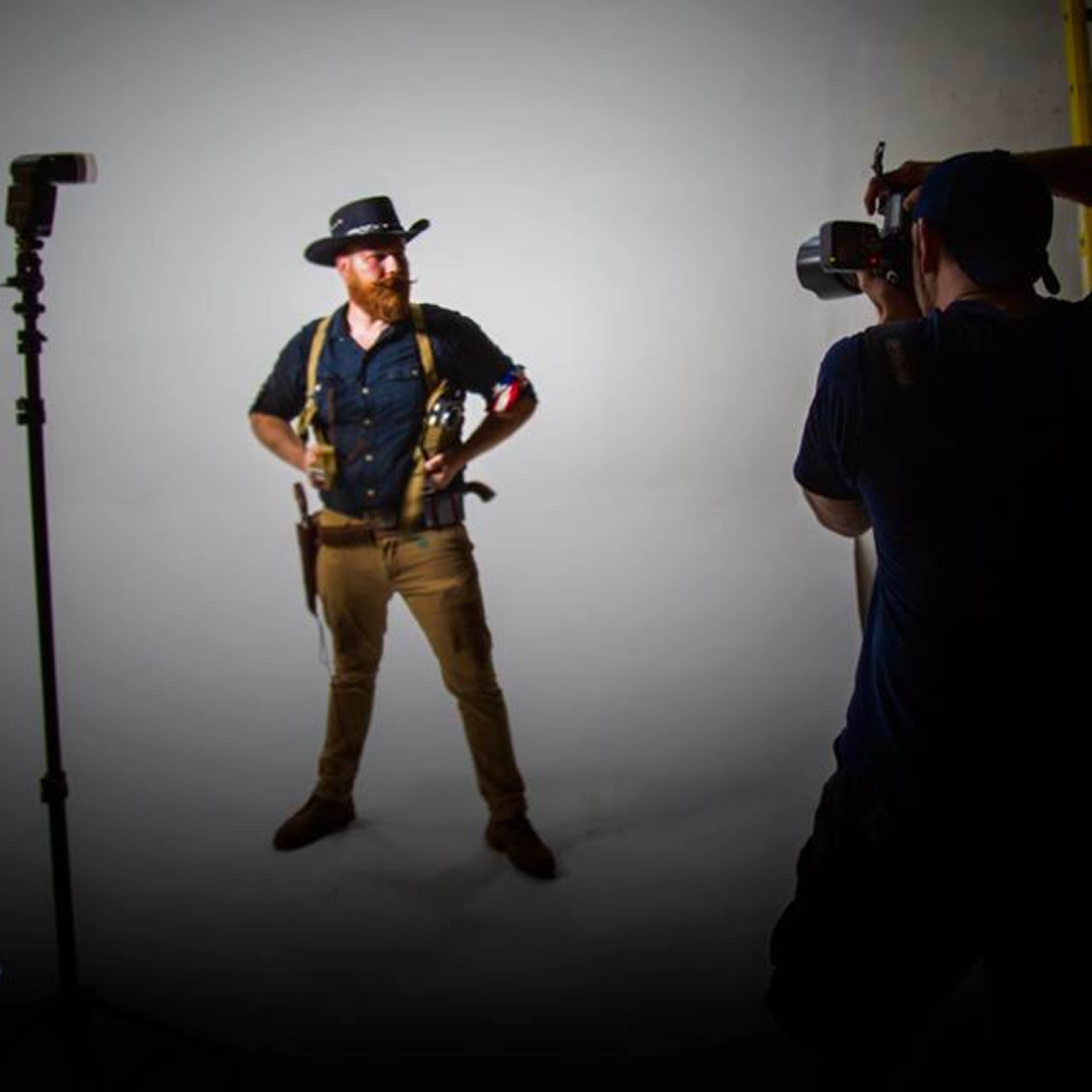 A man in a cowboy hat and gun holsters stands in front of a plain backdrop while a photographer captures his pose.