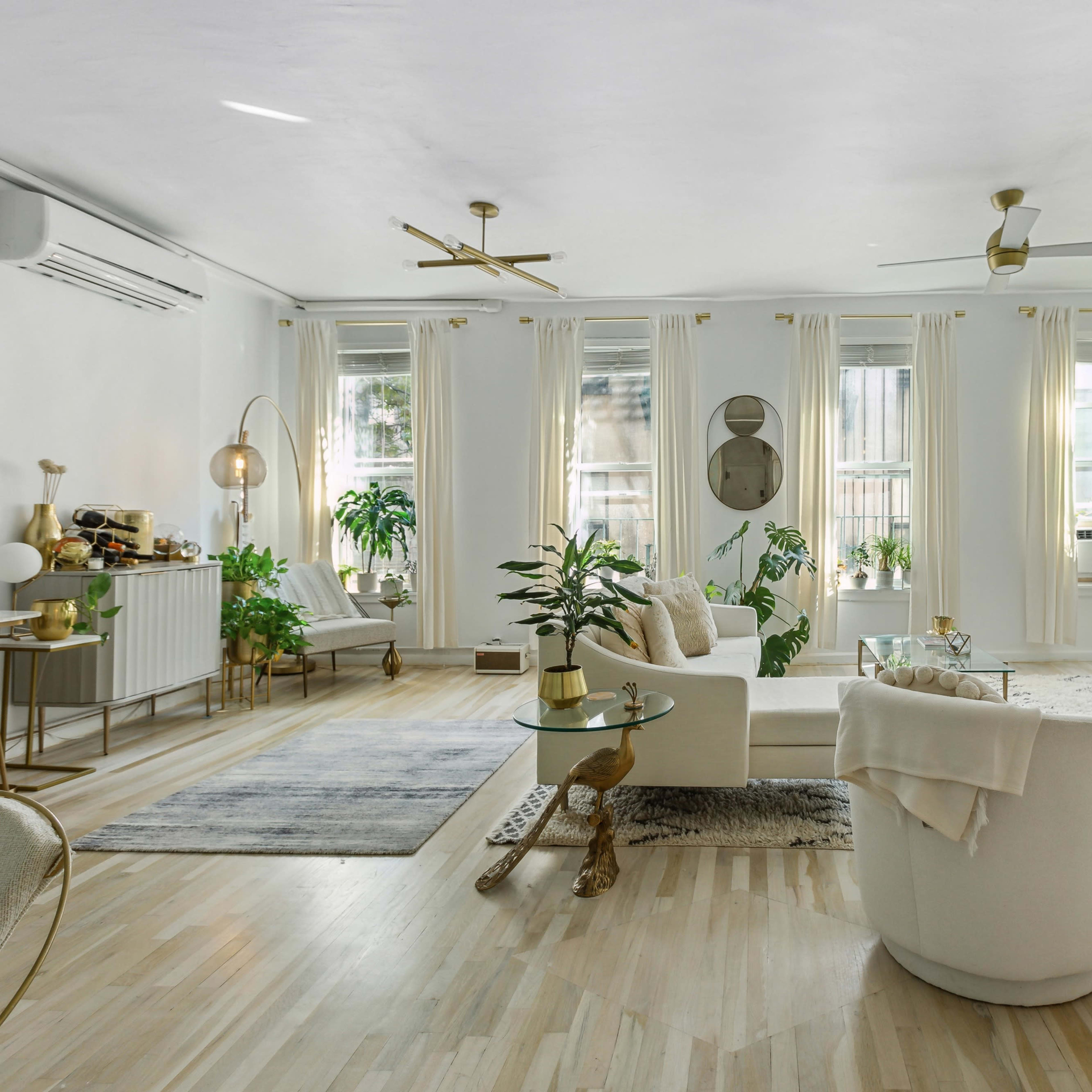 A bright living room features large windows, light wooden flooring, two white armchairs, a coffee table, and a wall-mounted television.