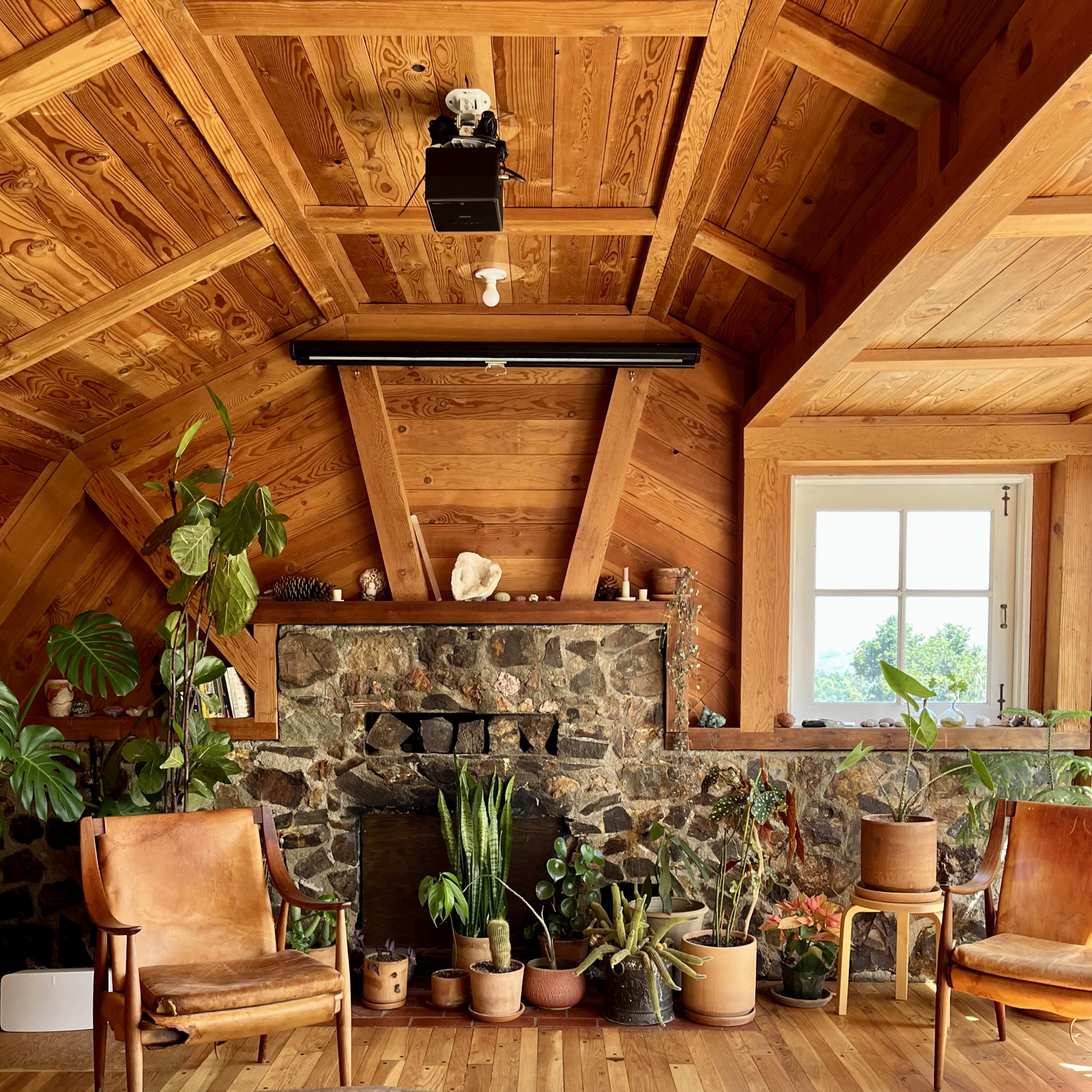 The interior of a wooden cabin features a stone fireplace, potted plants, two leather chairs, and a round wood coffee table.