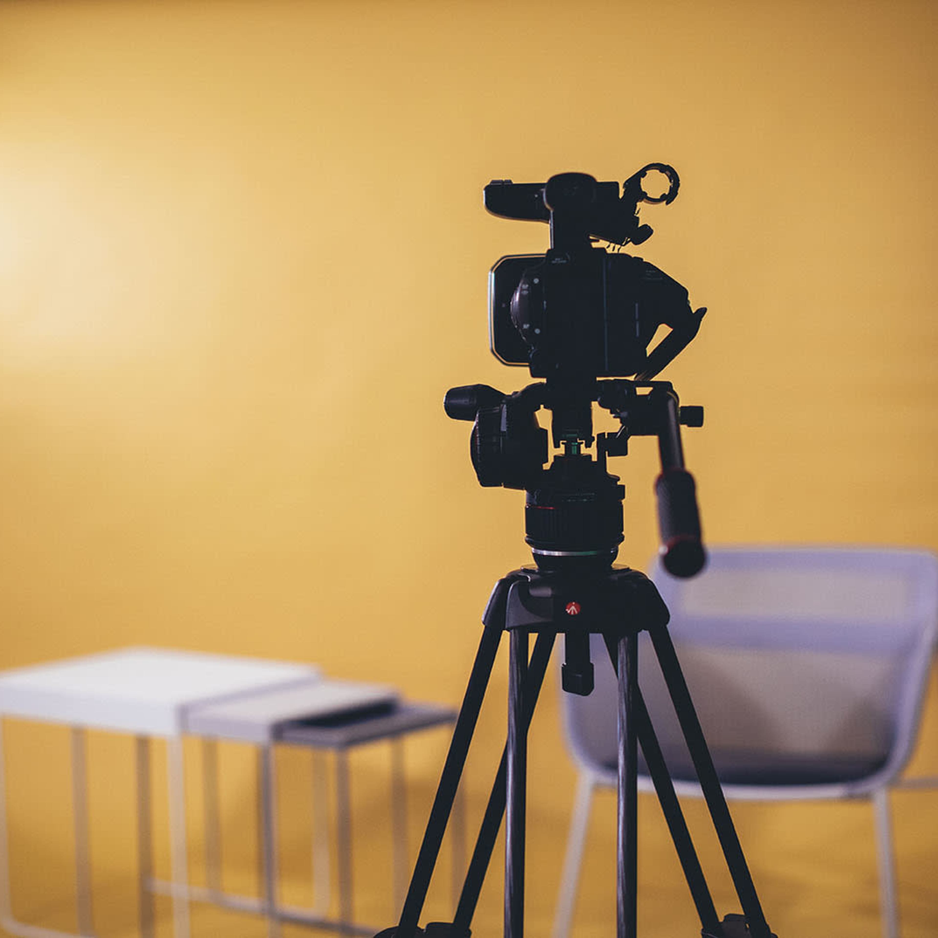 A camera on a tripod faces a yellow backdrop with a white table and a chair positioned nearby.