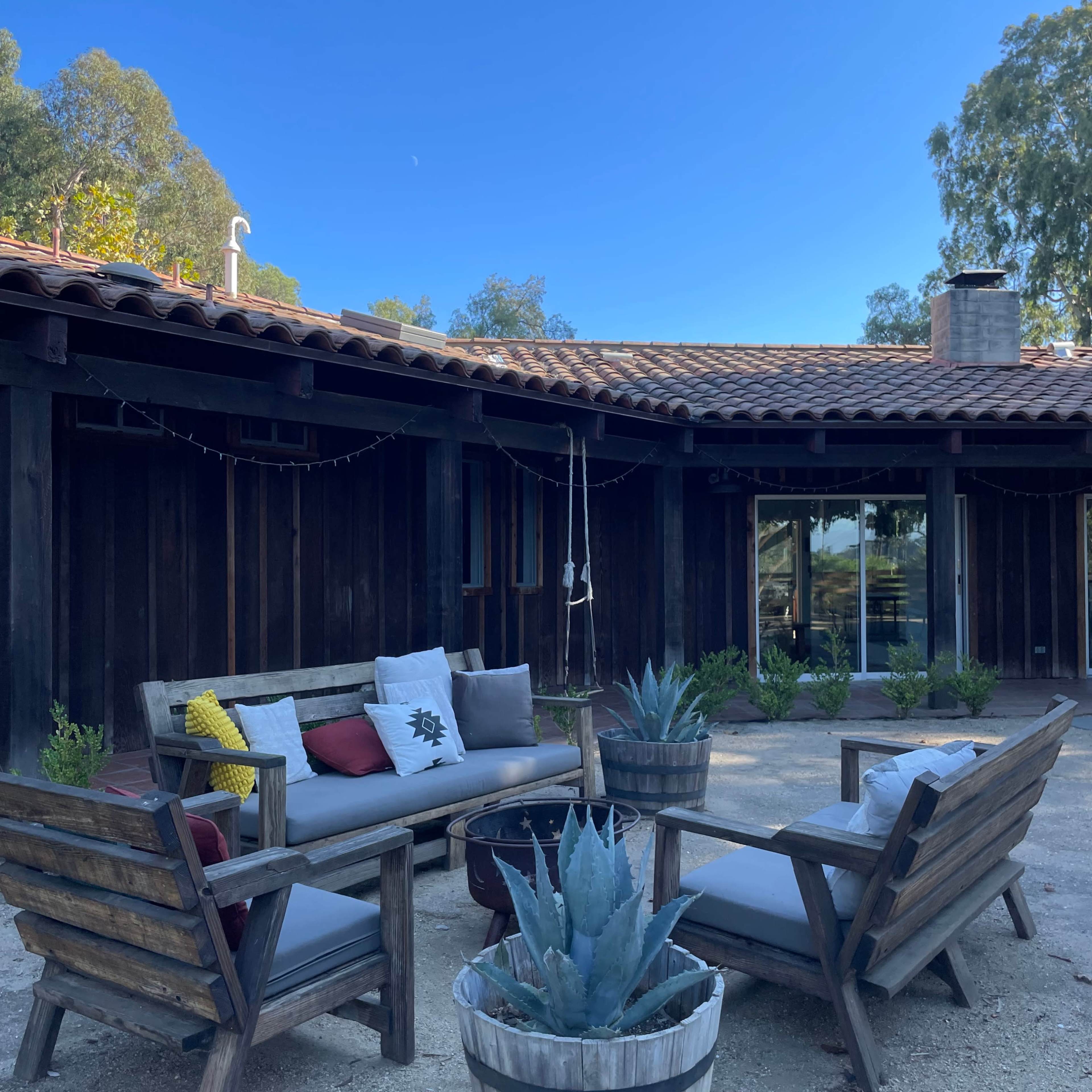 A cozy outdoor seating area features a sofa and two chairs arranged around a central planter filled with an agave plant, set in front of a rustic wooden building.
