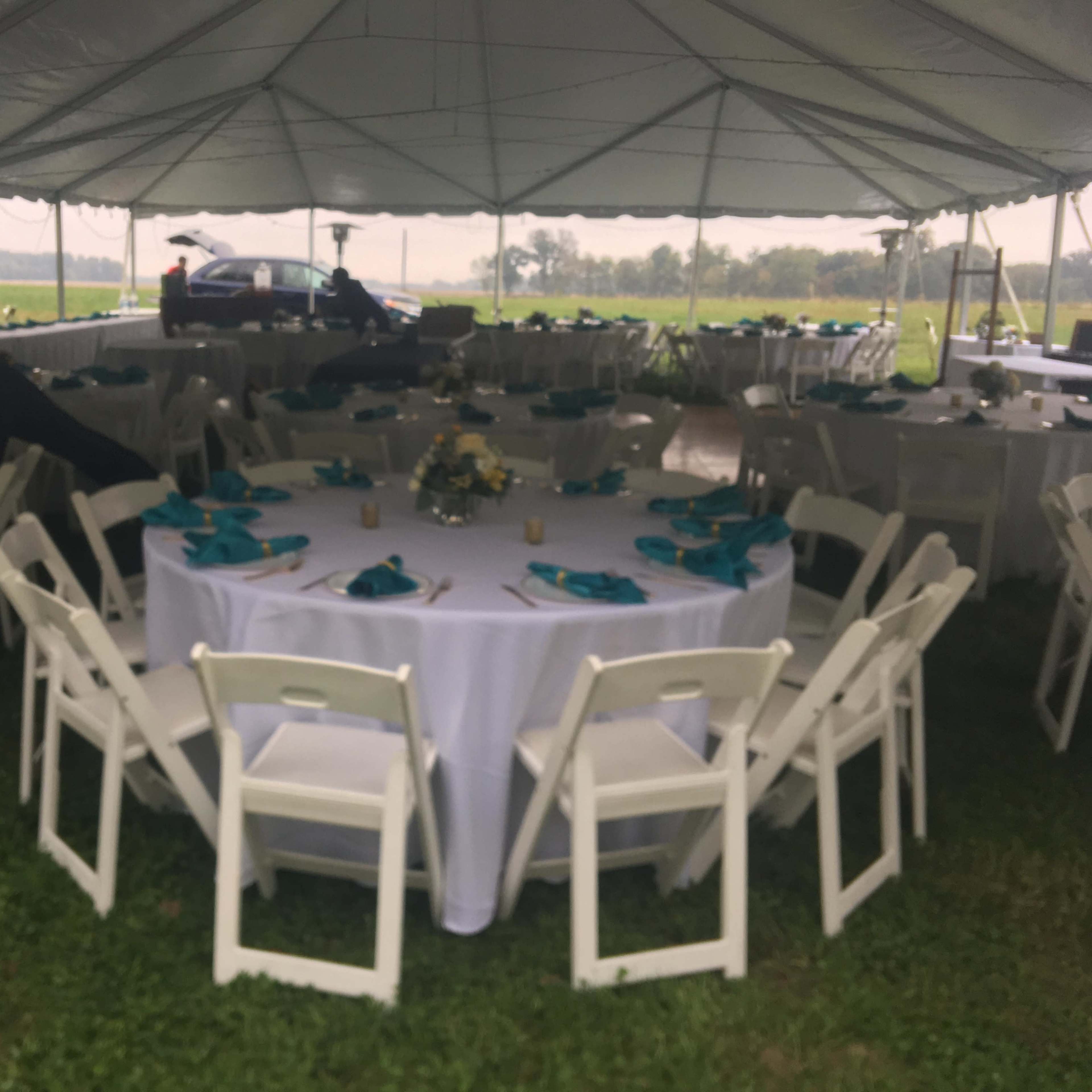 A spacious tent with neatly arranged round tables covered in white tablecloths and teal napkins, surrounded by chairs, set up for an outdoor event.