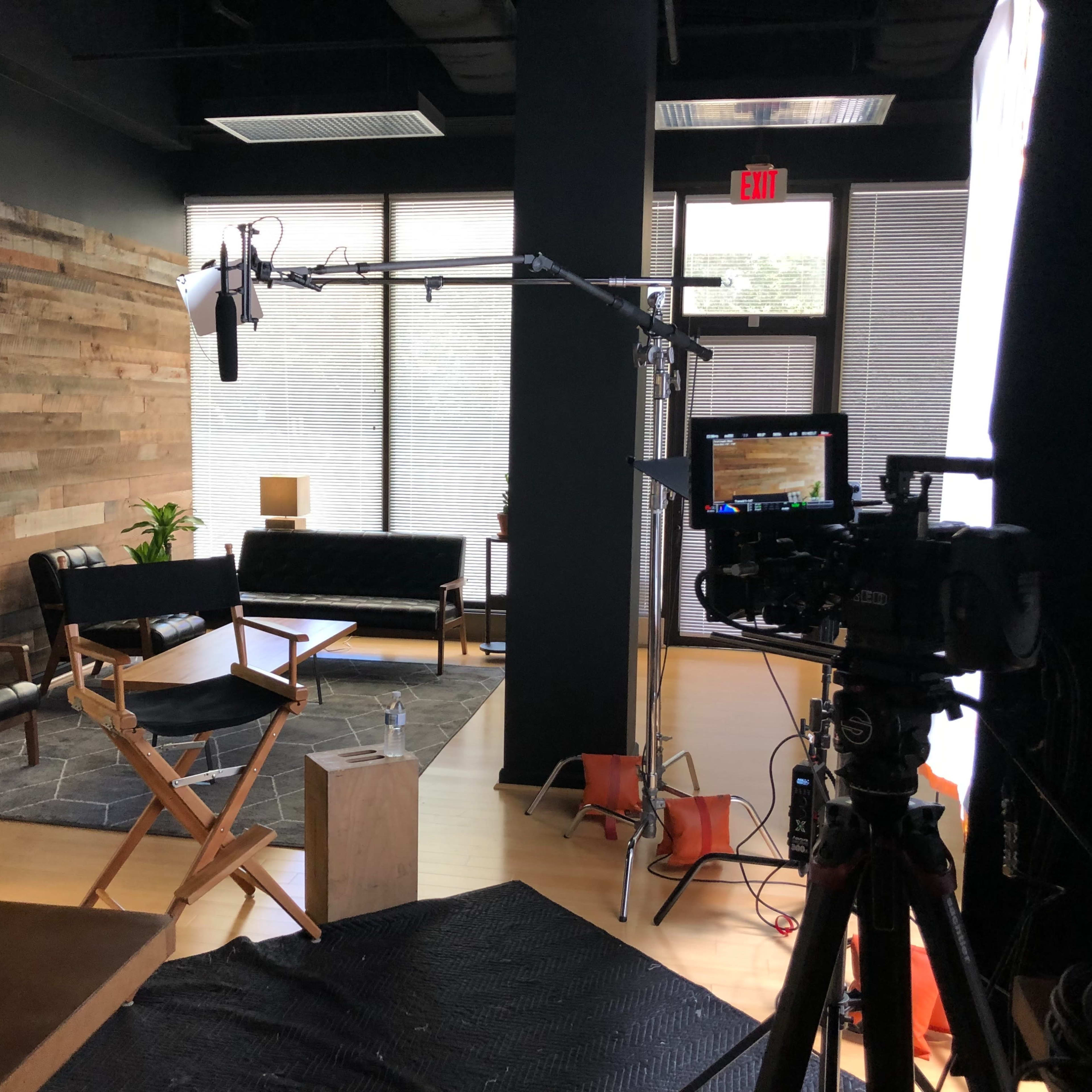 A film crew's setup is shown in a room featuring wooden paneling, a camera, lighting equipment, and various pieces of furniture.