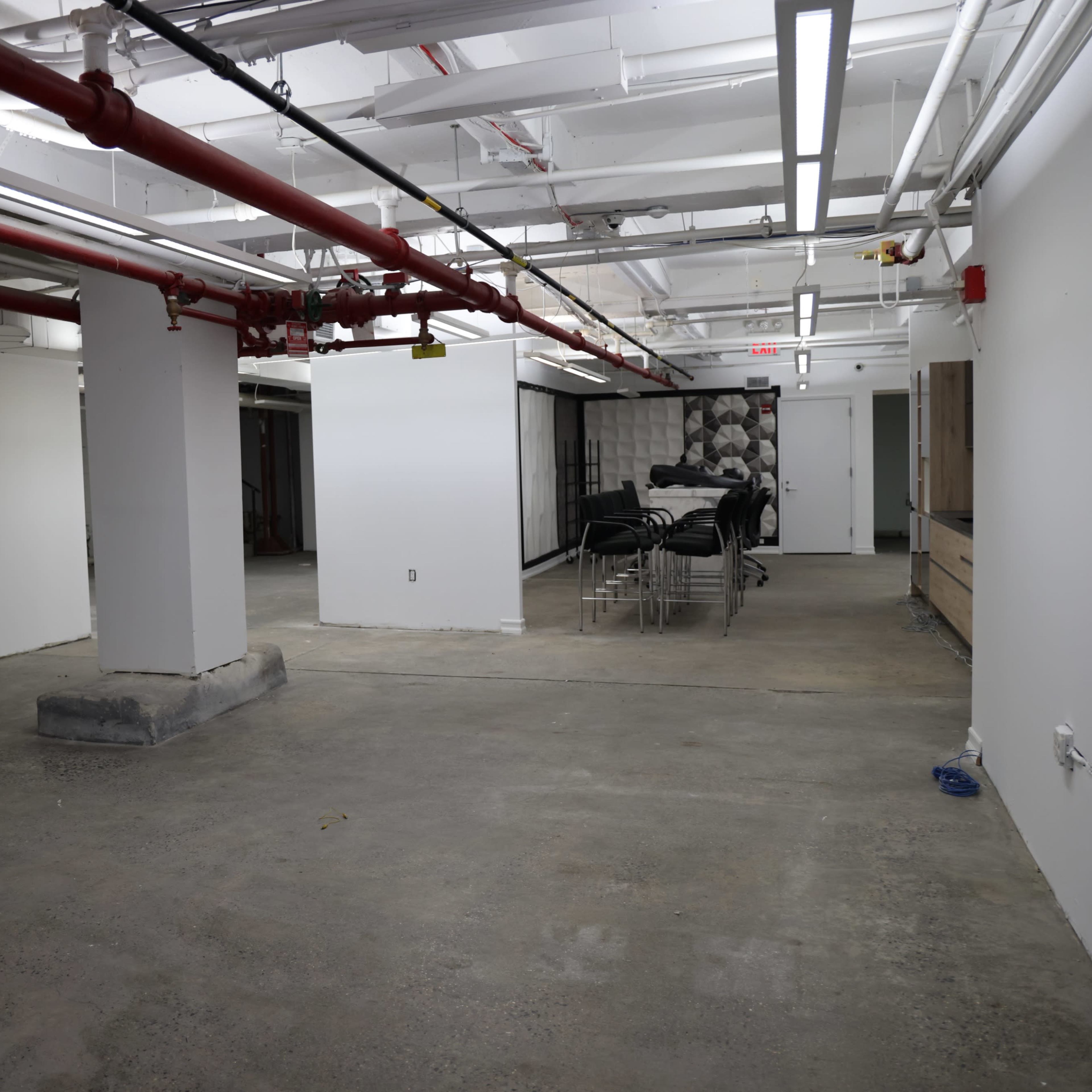 An empty, unfinished commercial space with exposed ceilings, concrete flooring, and a stack of chairs against one wall.