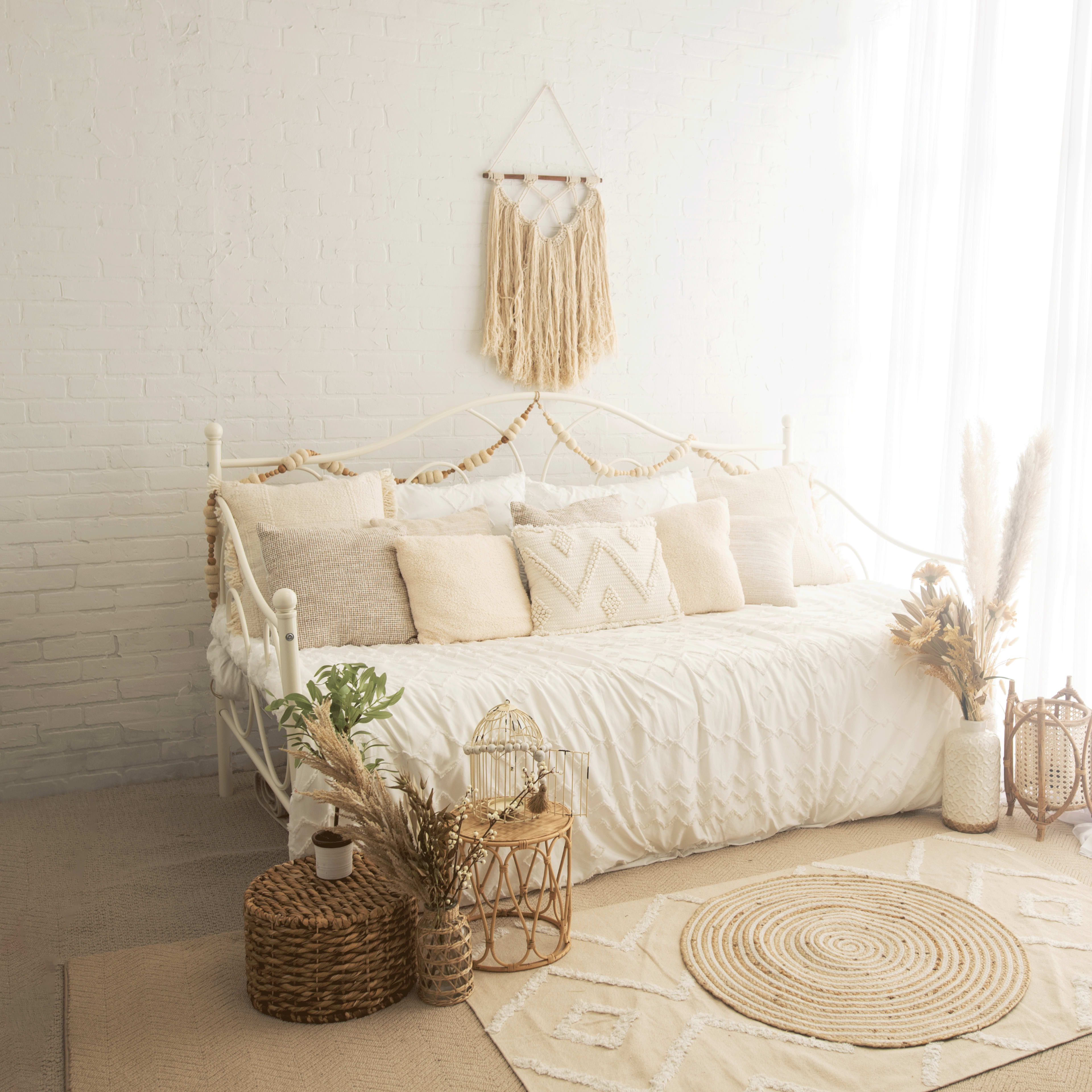 A bright, minimalist bedroom featuring a white daybed adorned with decorative pillows, a woven rug, and natural elements like plants and baskets.