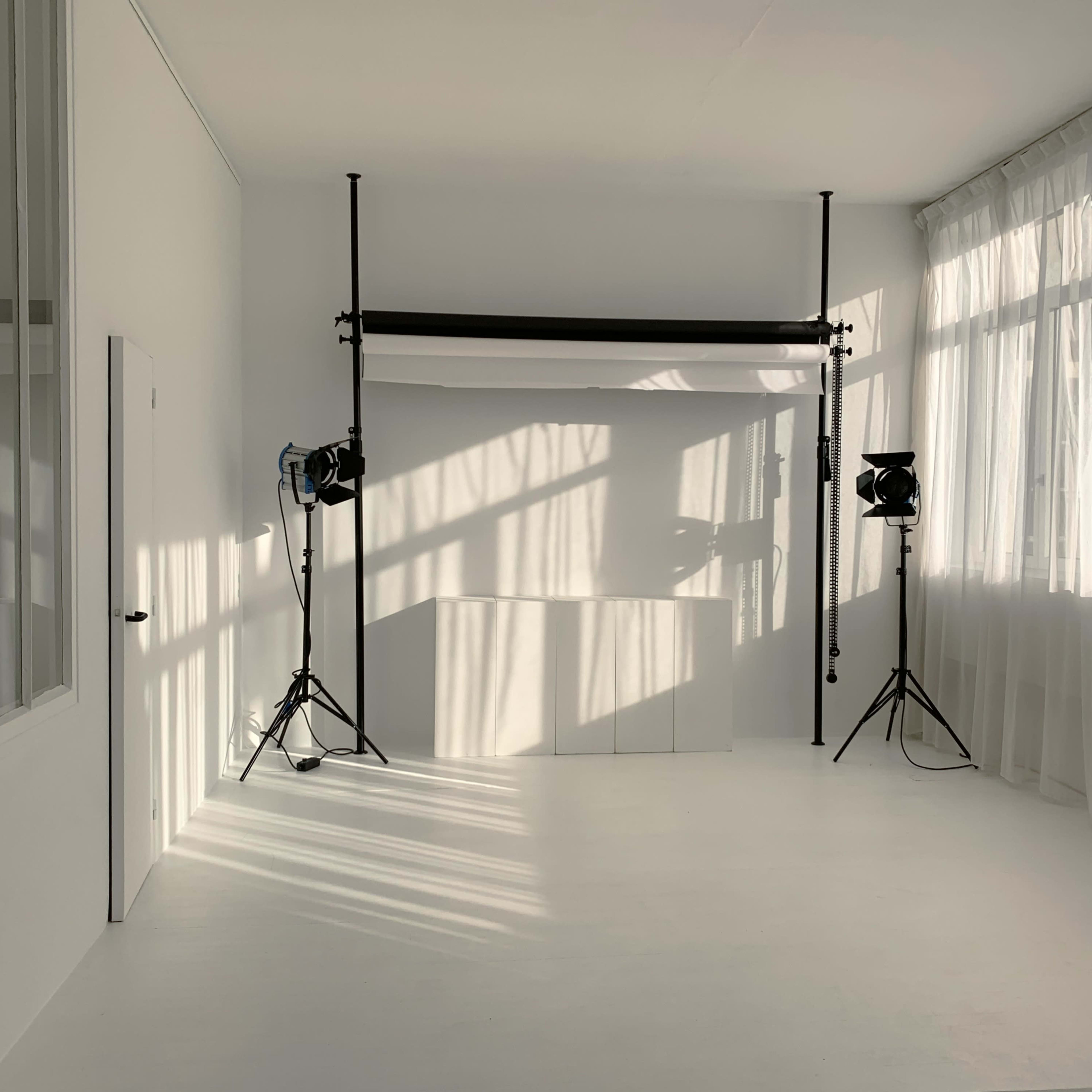 A bright photography studio with white walls, a backdrop stand, and two light stands casting shadows on the floor.