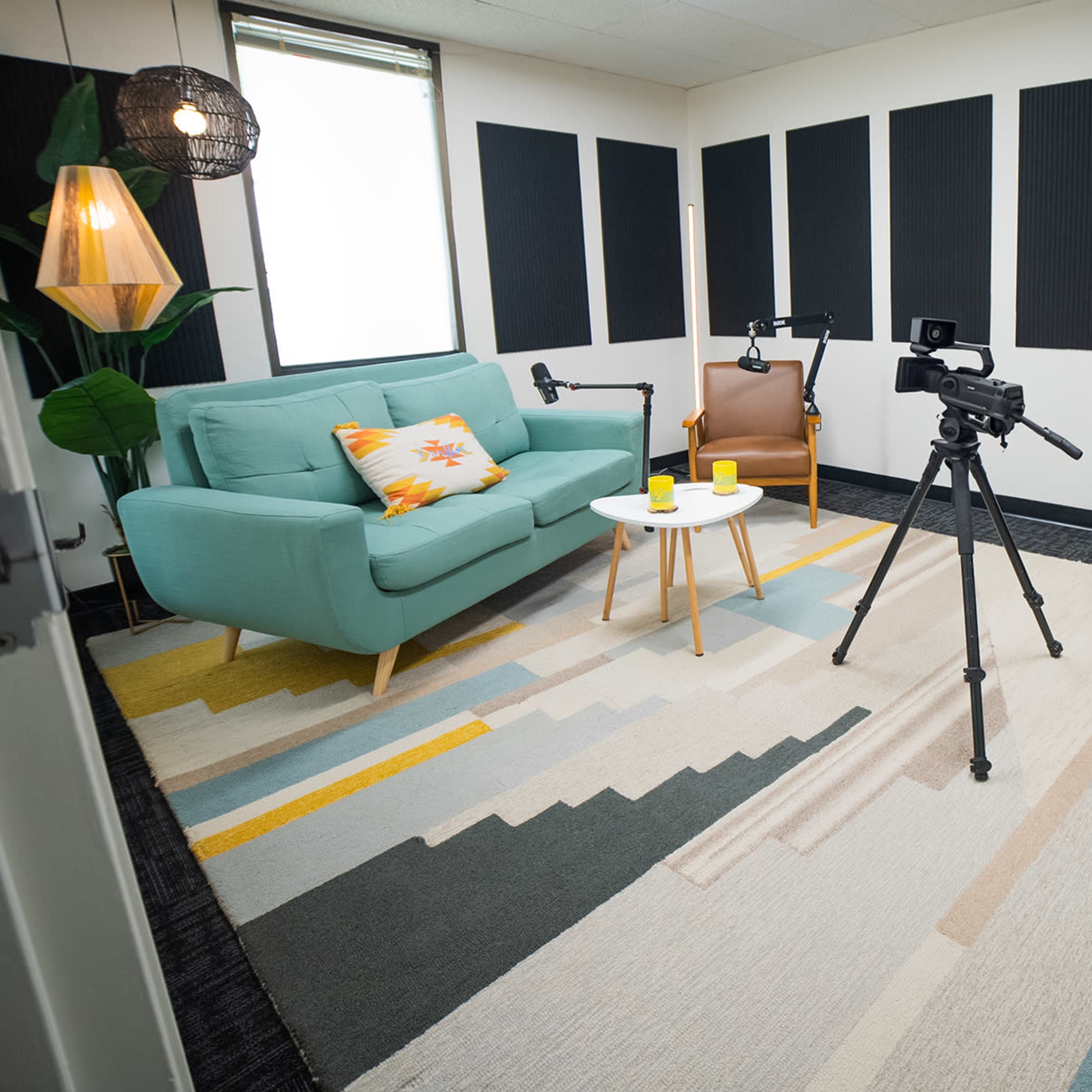 A modern studio space with a teal couch, a small table, a camera on a tripod, and a soundproof booth in the background.