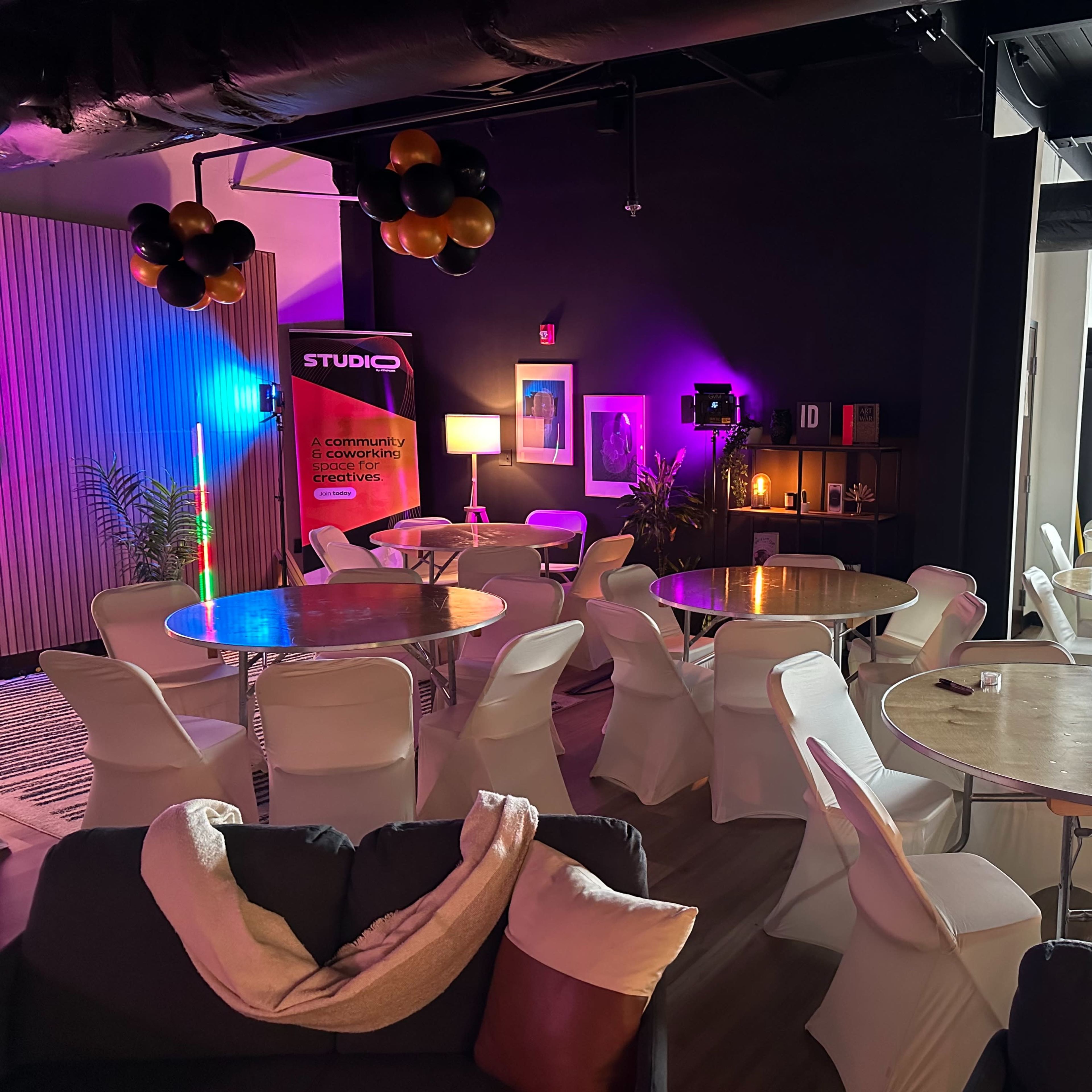A modern event space with round tables and chairs arranged for a gathering, decorated with balloons and colorful lighting.
