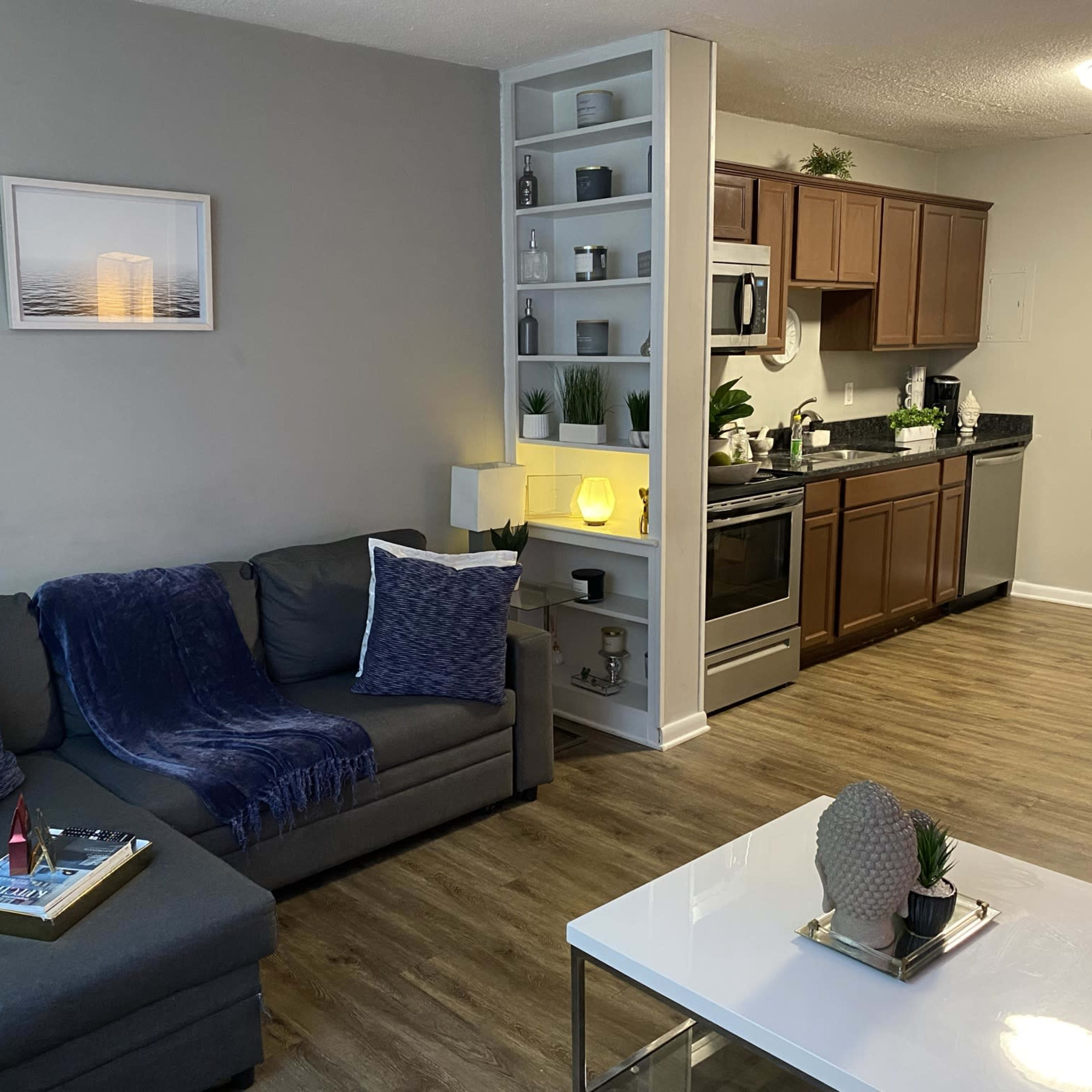 A modern living room with a gray sofa, a white coffee table, a kitchen in the background, and decorative shelving featuring plants and a light source.