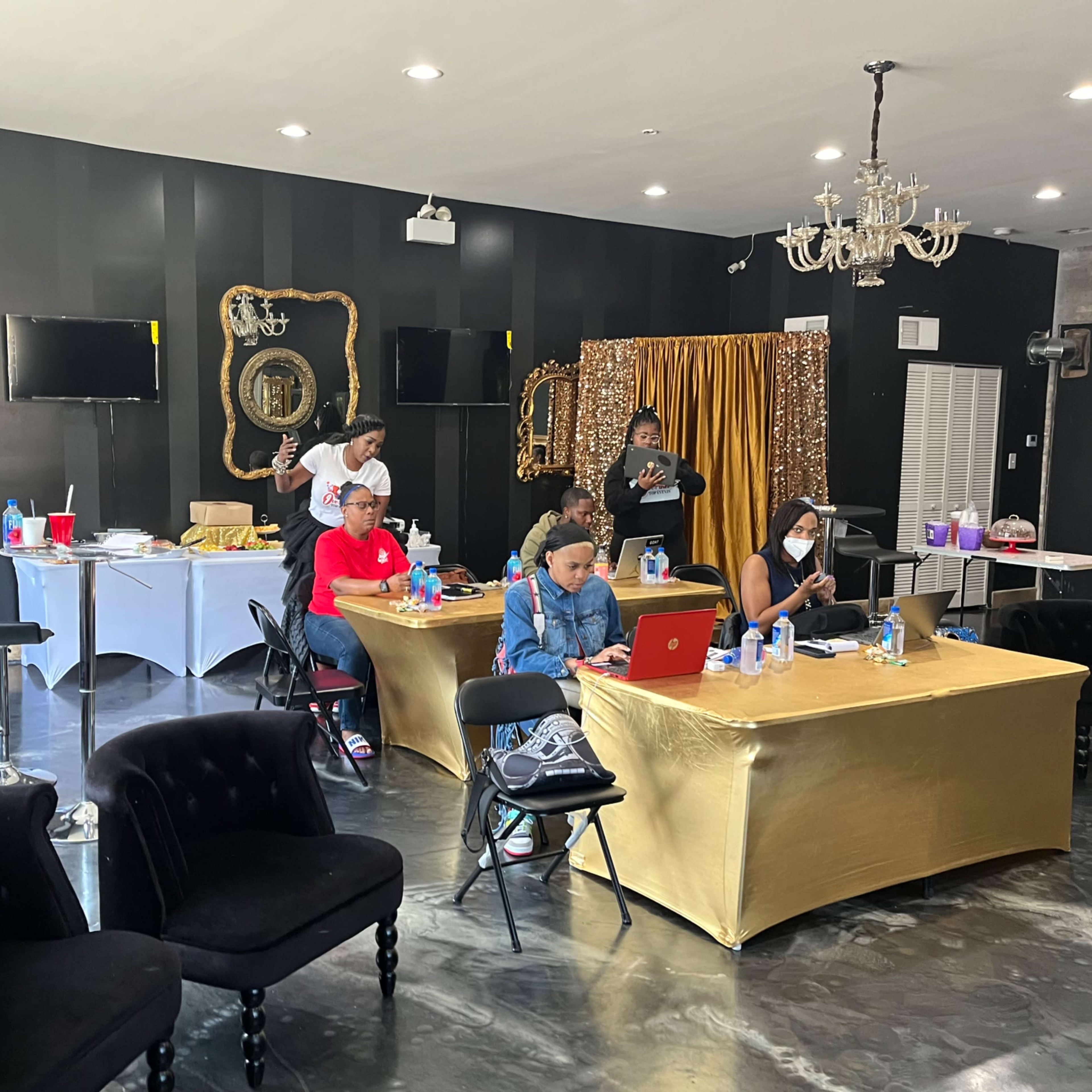 A group of people is gathered in a room with black walls and elegant decorations, working on laptops while some prepare food at a table in the background.