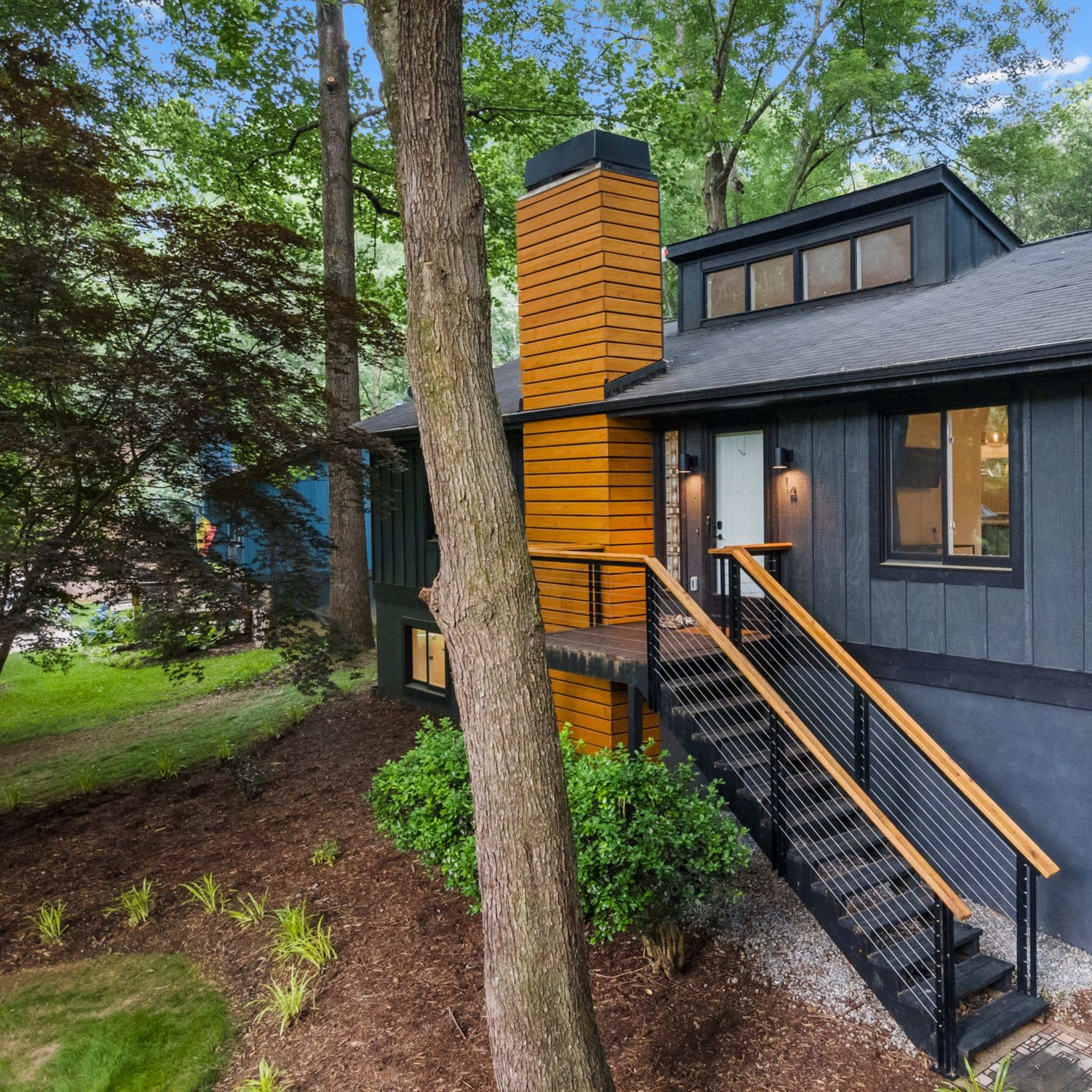 A modern black and wood-paneled house with a sloped roof and a chimney, surrounded by trees and landscaped greenery.