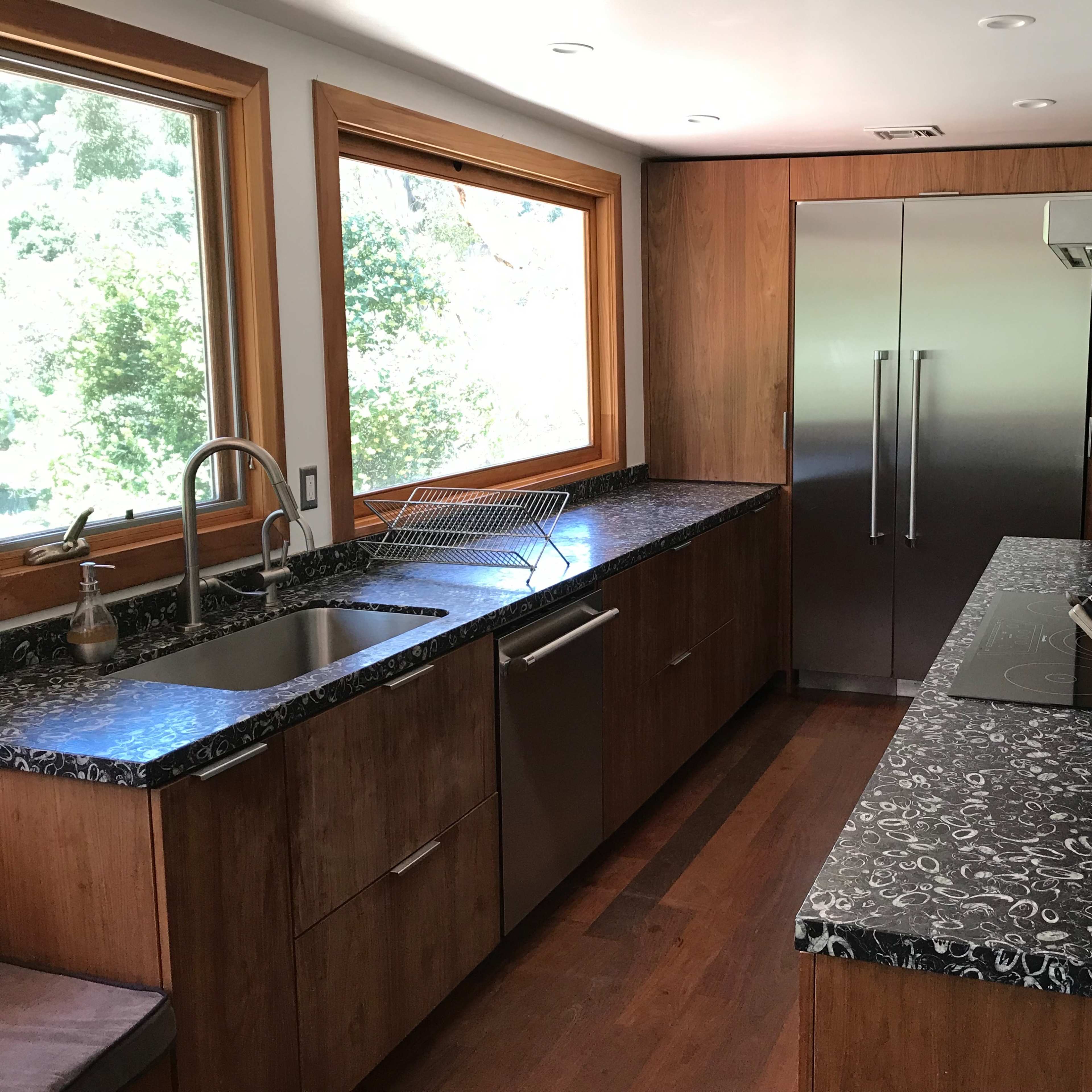 A modern kitchen features a sink, stainless steel refrigerator, and dark patterned countertops with wooden cabinetry and natural light coming through large windows.