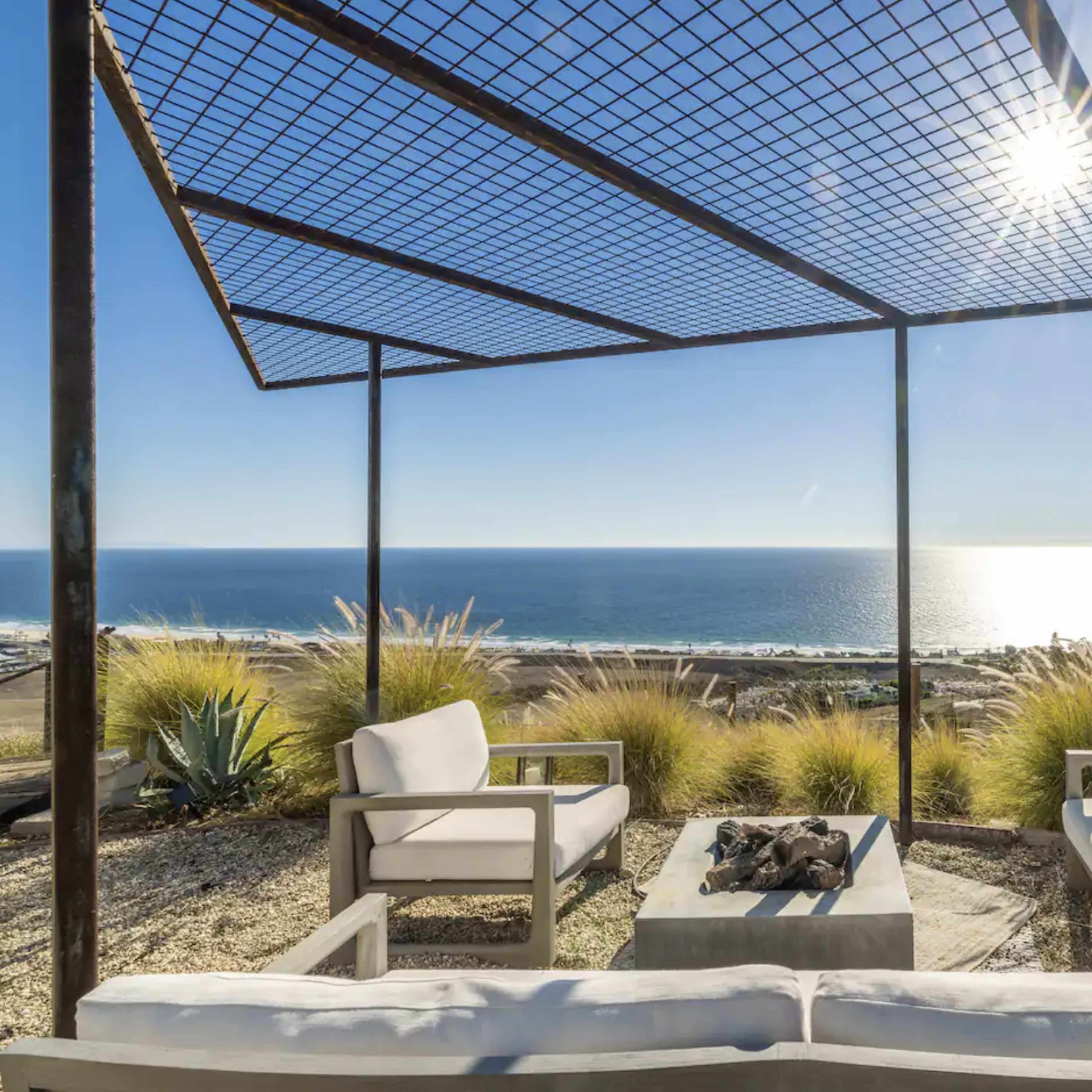 A modern outdoor seating area features white armchairs and a stone table around a fire pit, overlooking the ocean under a lattice canopy.
