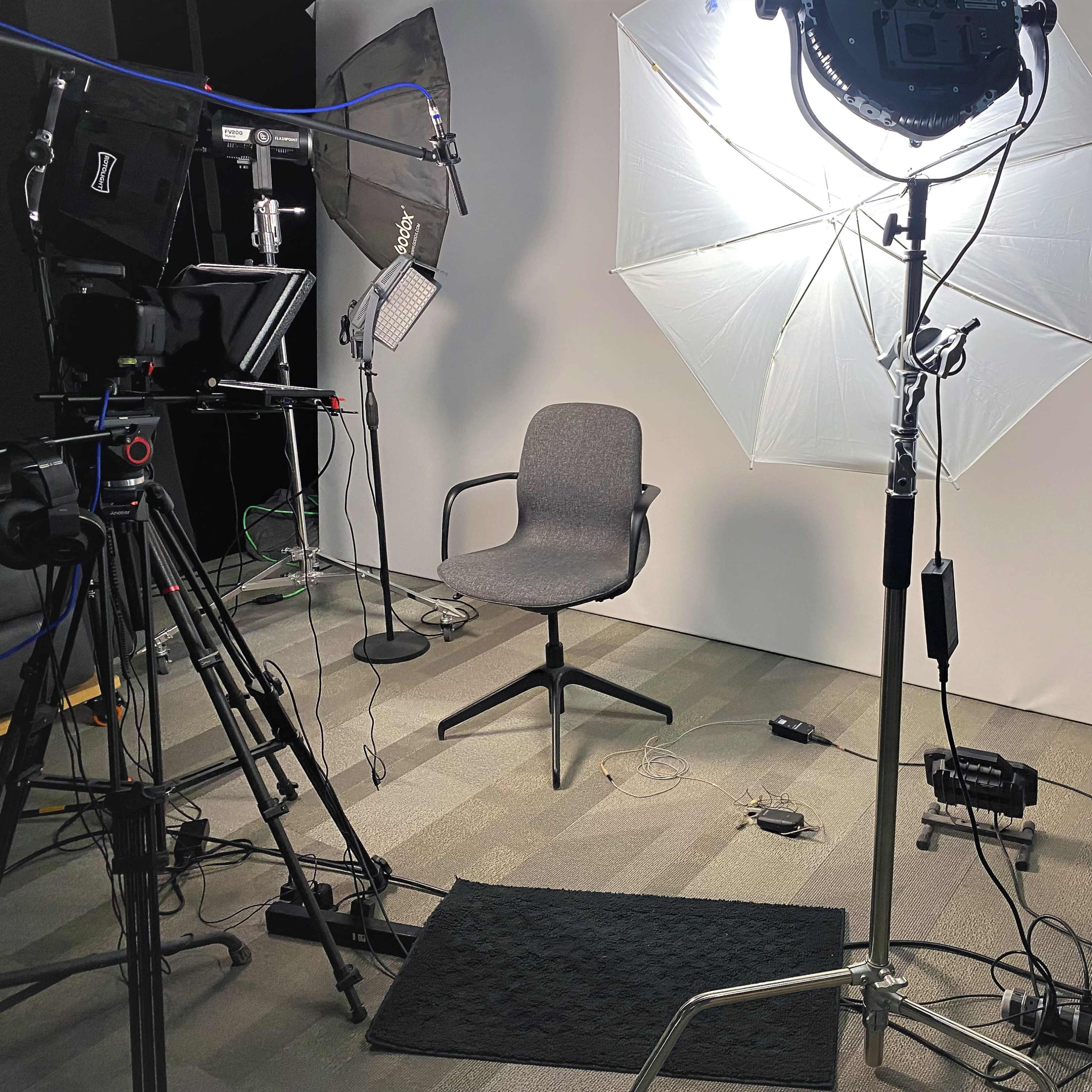 A filming setup with a gray chair under softbox lights, surrounded by camera equipment and a backdrop.