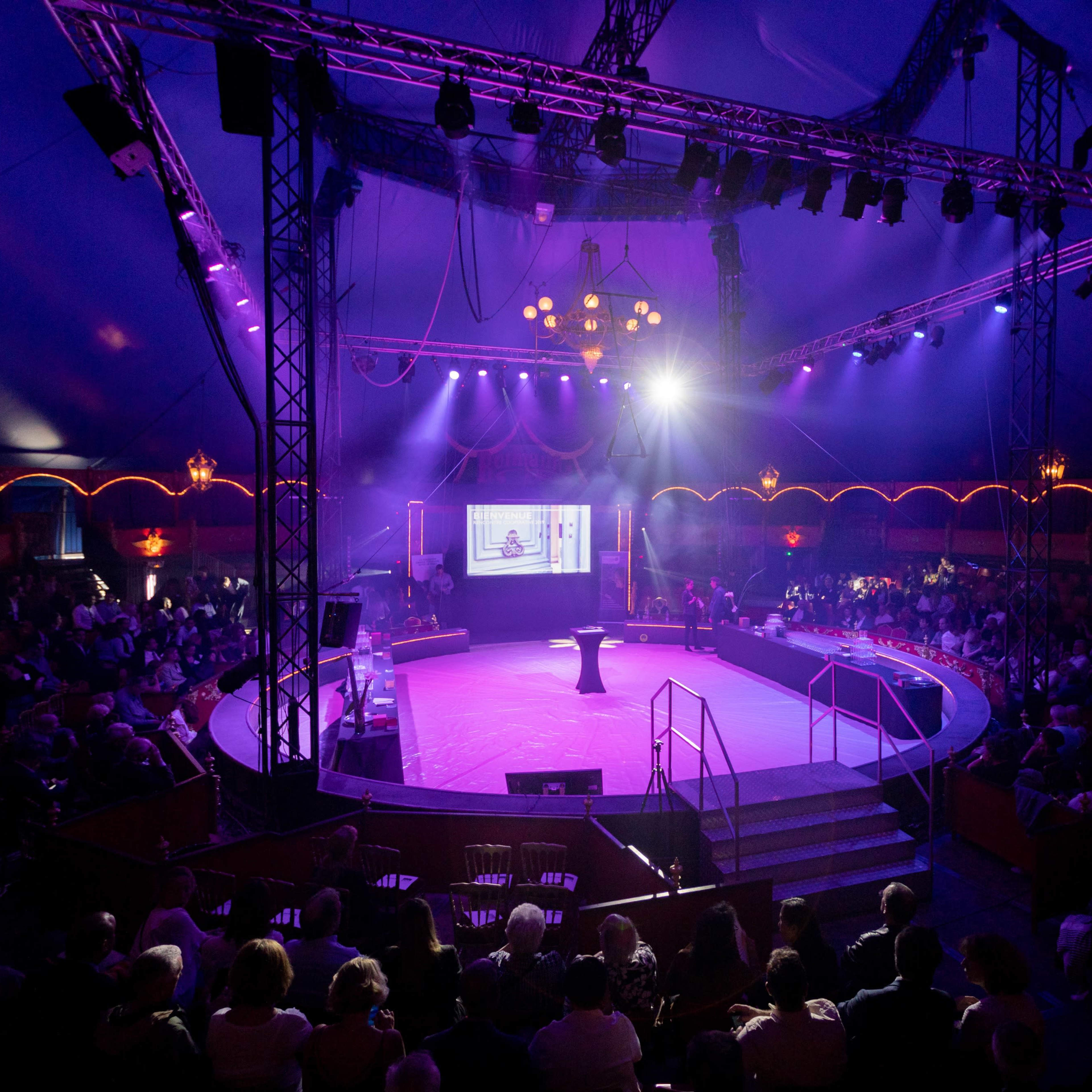 A crowd gathers under a large circus tent with purple lighting, centered around a circular stage.