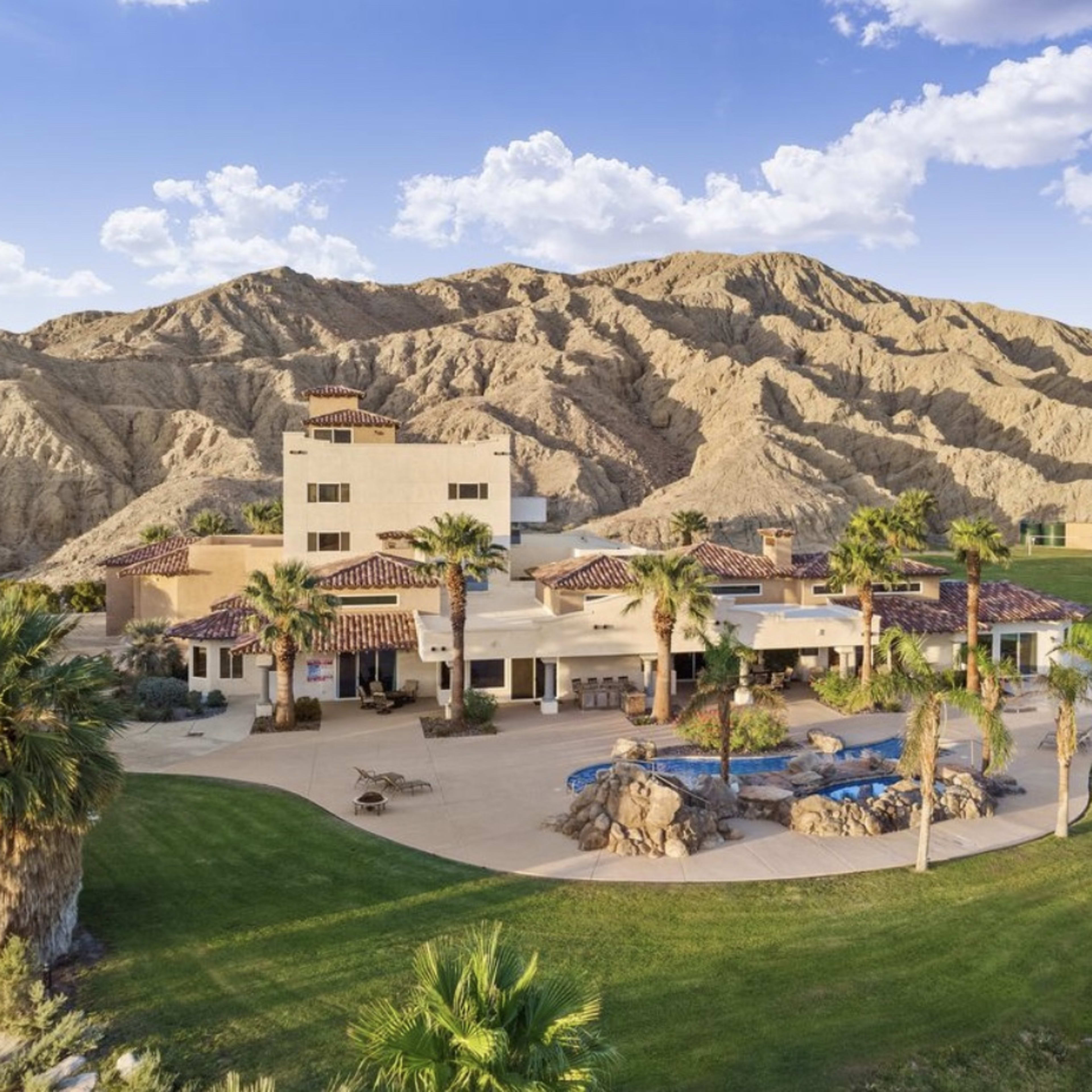 A spacious, modern home surrounded by lush greenery and desert-like hills in the background.