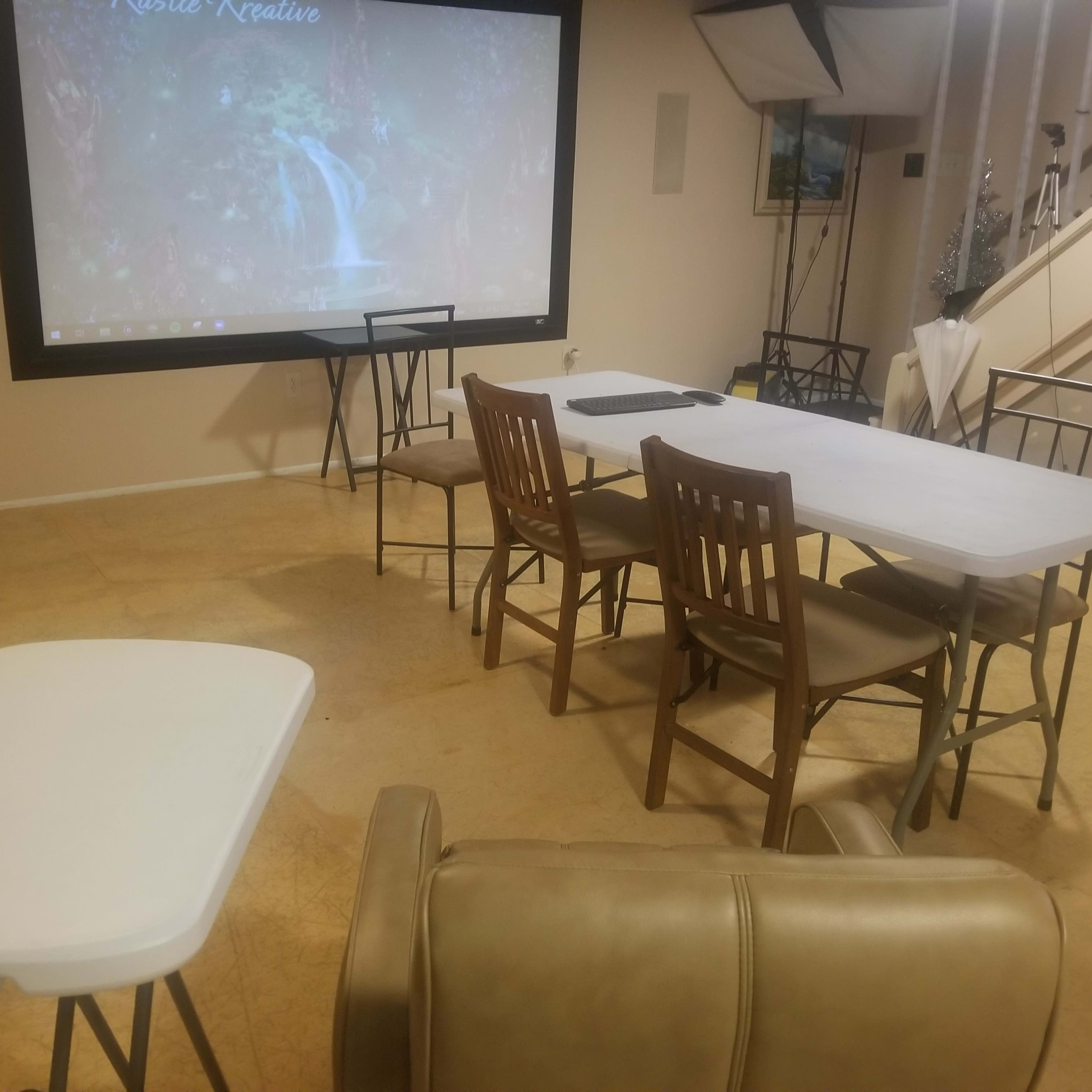 A small indoor room features a rectangular table surrounded by chairs, with a large screen displaying a nature scene on one wall.