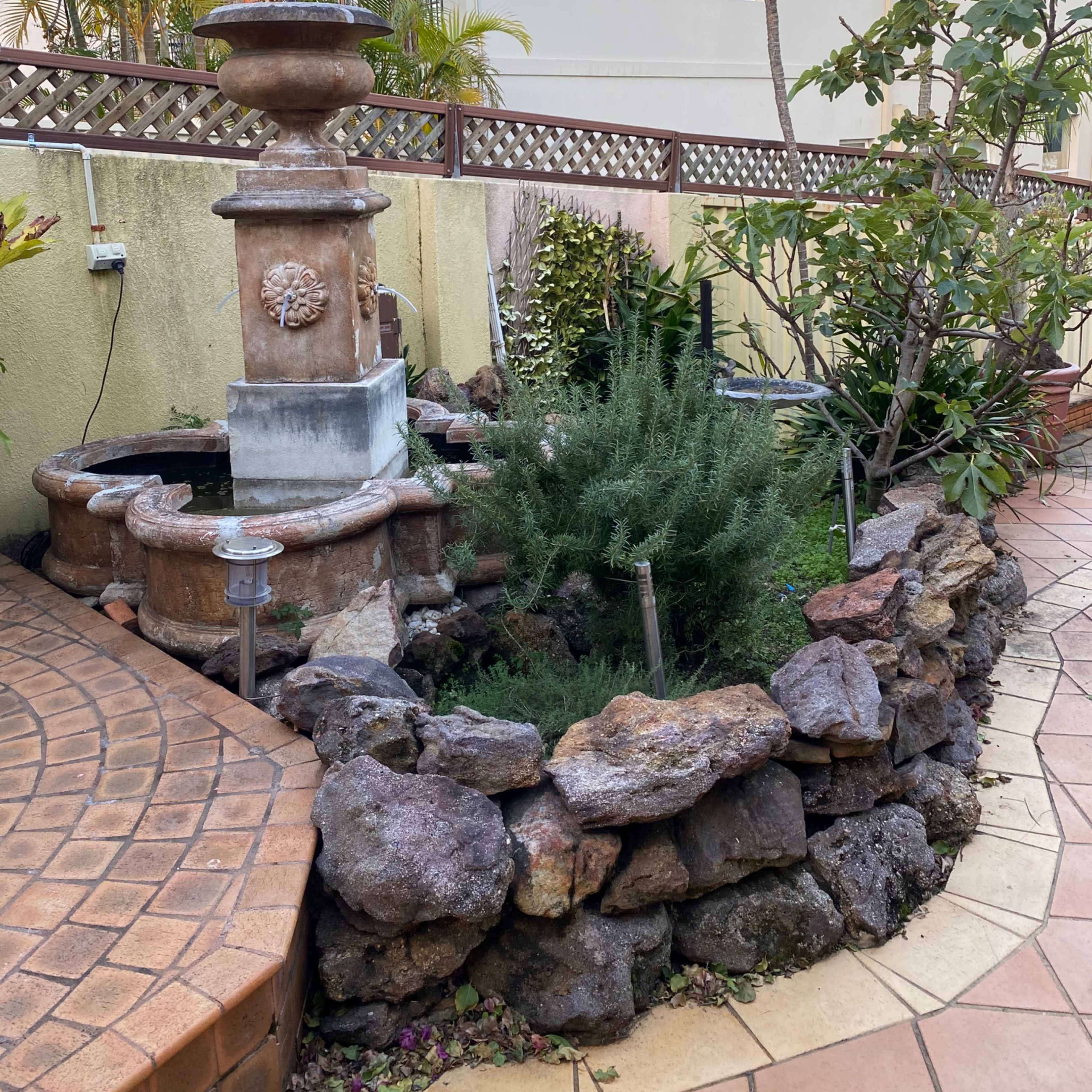 A stone-bordered garden area featuring a decorative fountain surrounded by greenery and tropical plants.