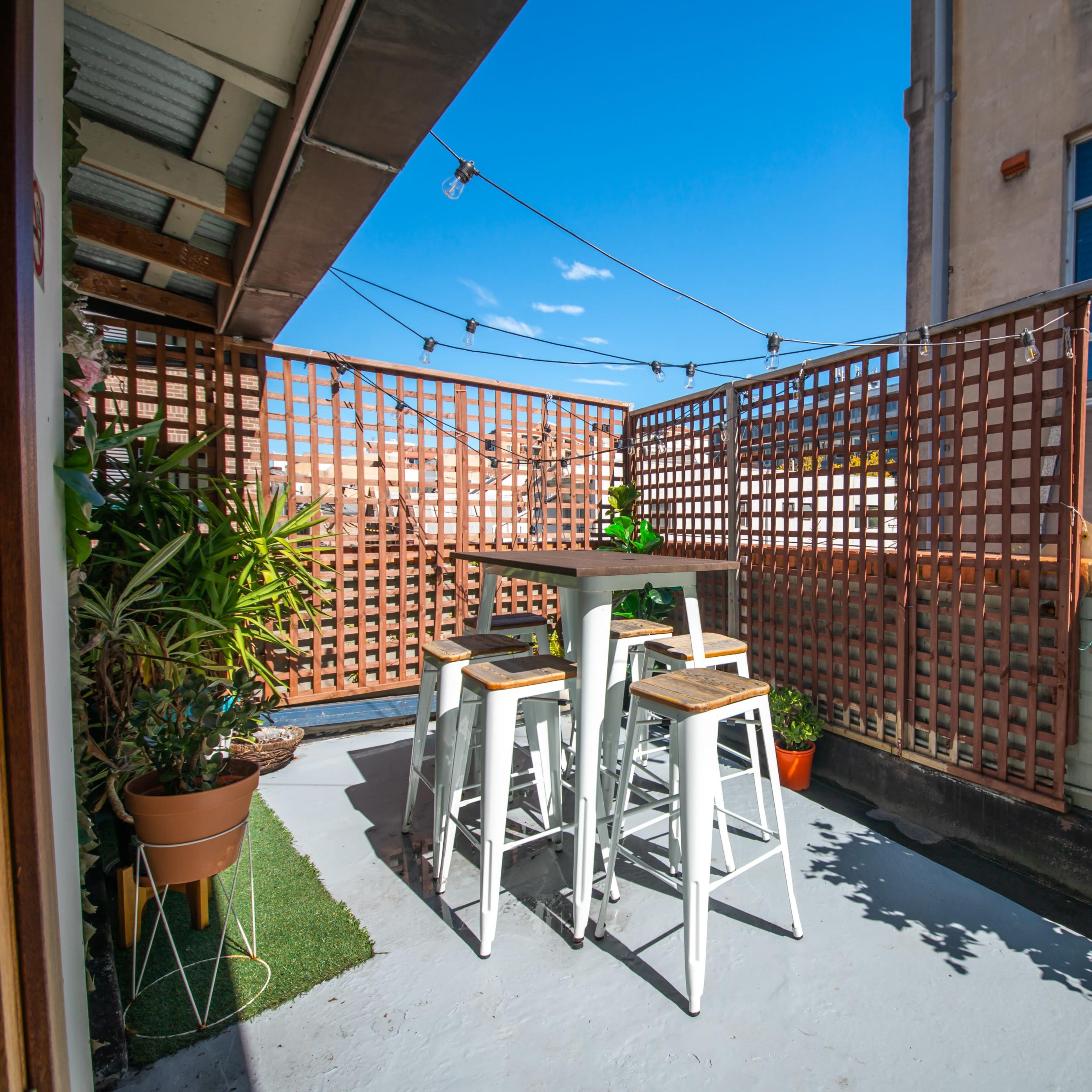 A small outdoor patio features a table with tall stools, surrounded by lattice fencing and potted plants.