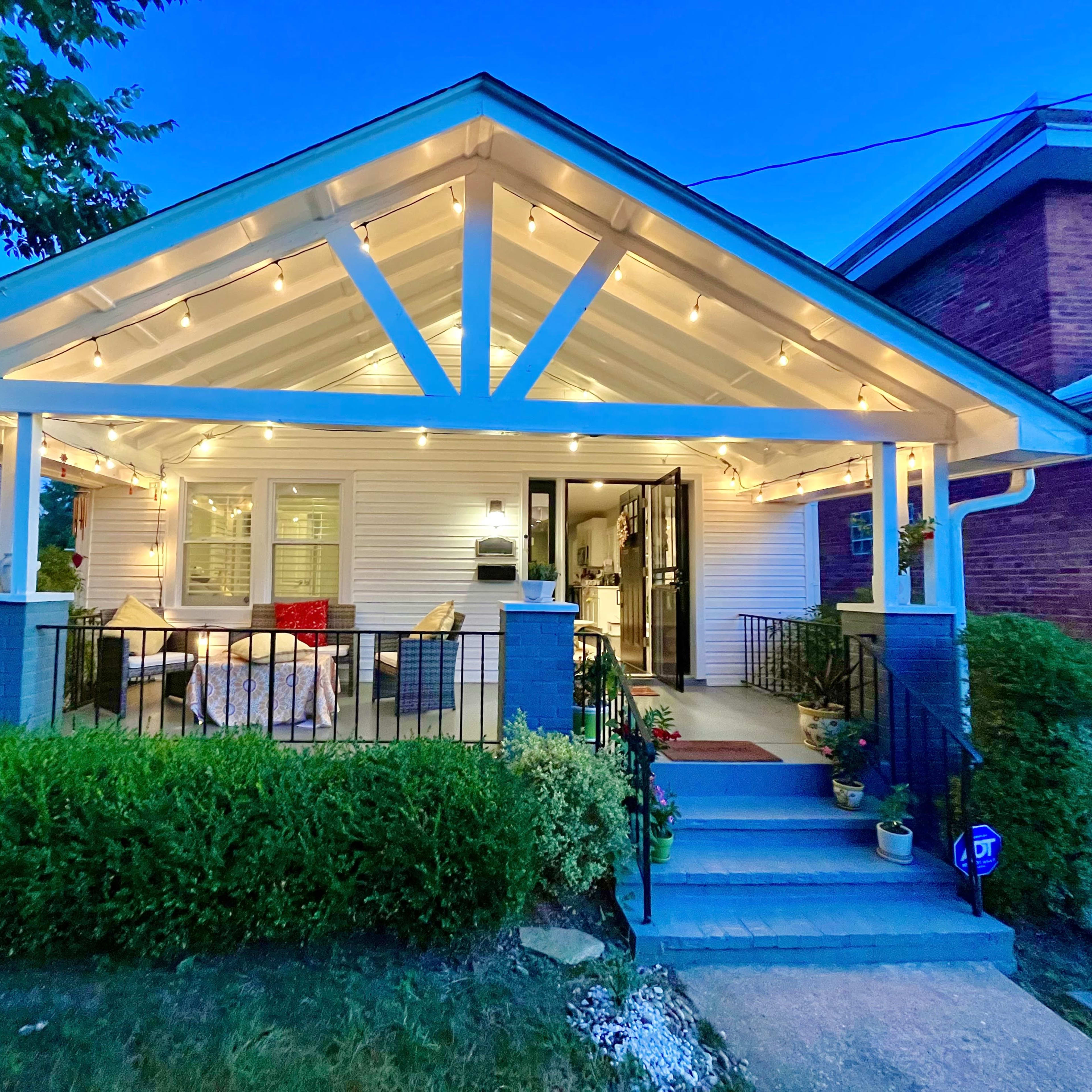 A well-lit porch of a house with decorative lights, surrounded by greenery and a welcoming entrance.