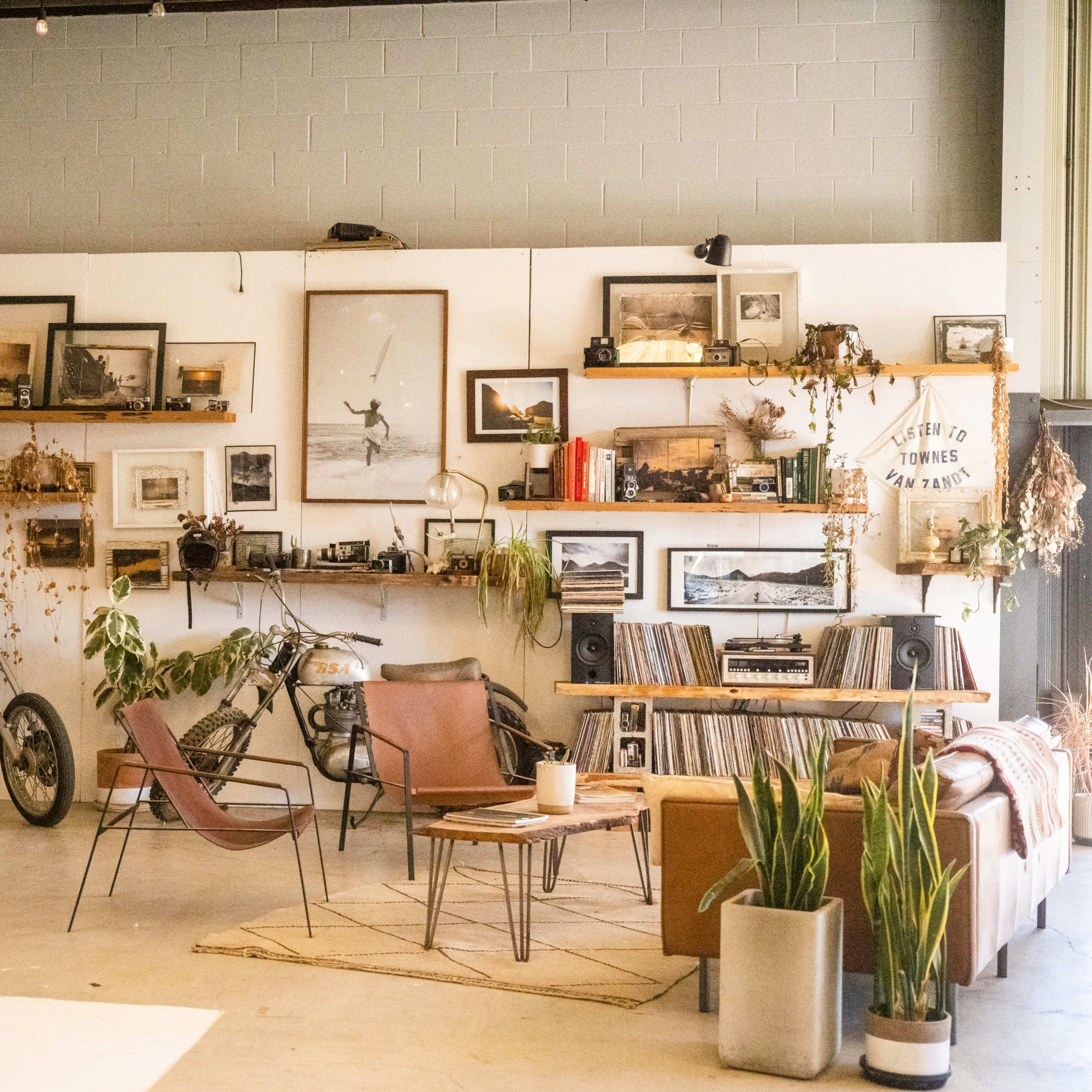 A spacious interior decorated with vintage photographs, plants, and motorcycle-themed decor, featuring a sofa, armchairs, and a record player.