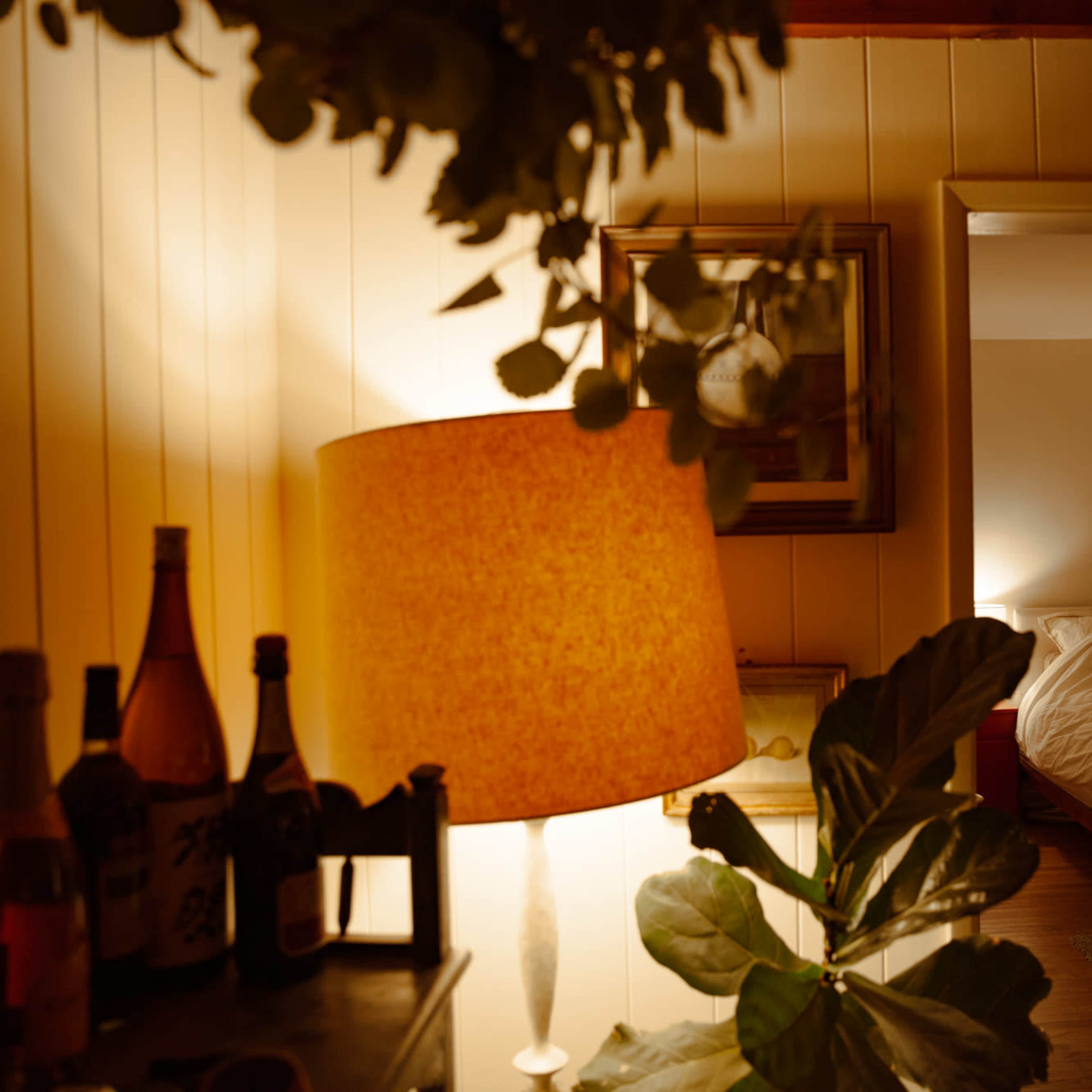 A warm-lit room with a lamp, bottles on a table, a potted plant, and a doorway leading to a bedroom with a unmade bed.