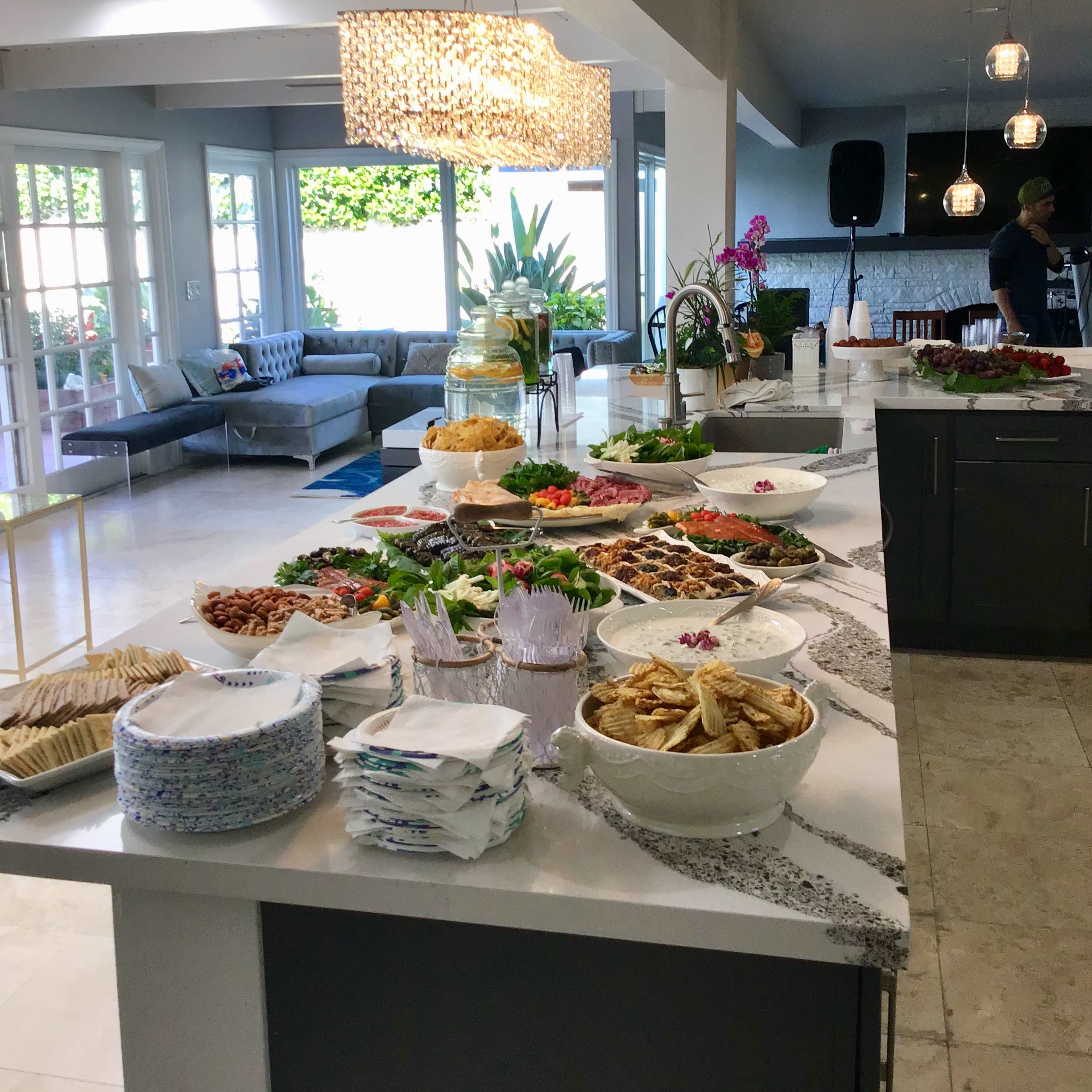 A long table is set with various food dishes, including salads, snacks, and desserts, in a well-lit modern kitchen and dining area.