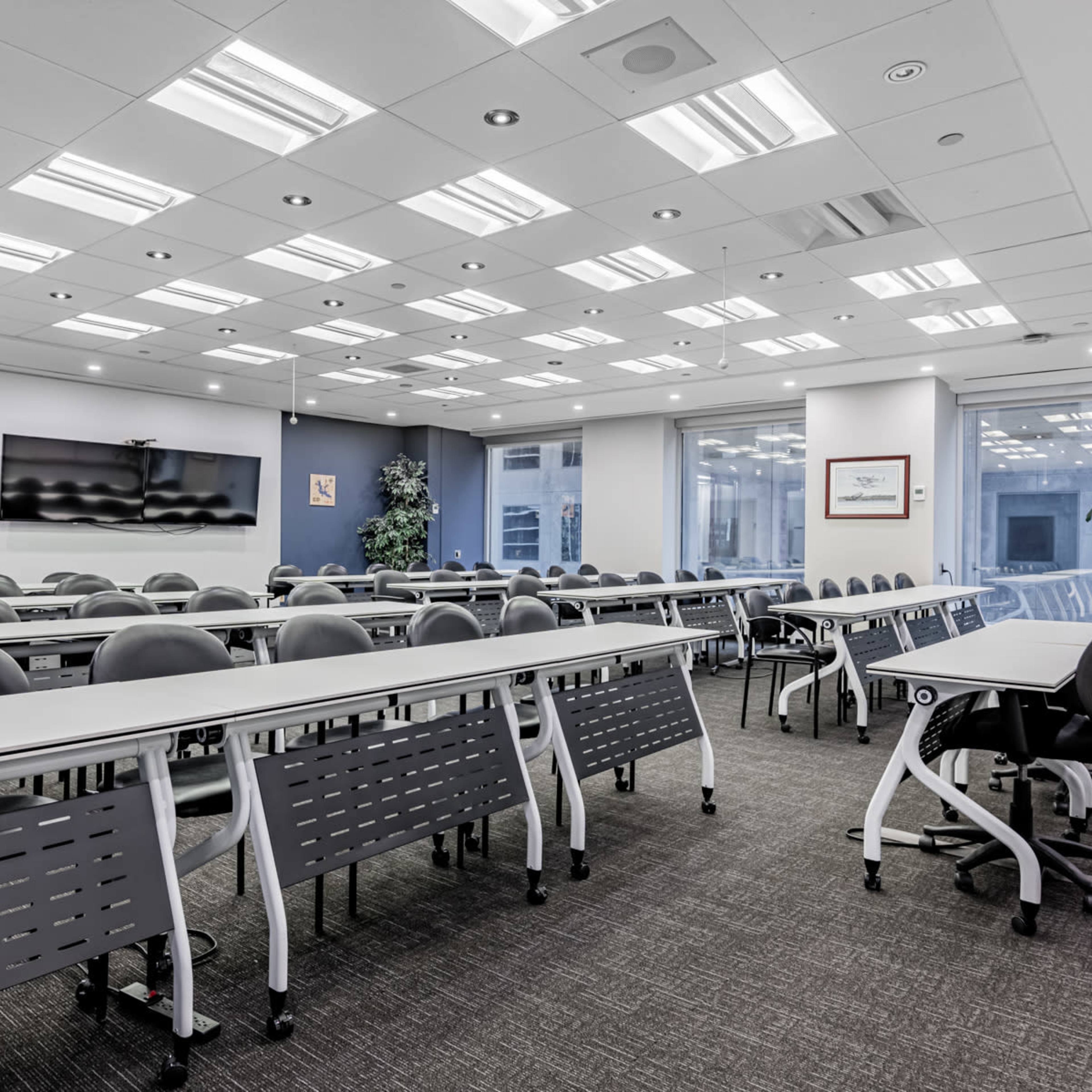 A modern conference room with rows of tables and chairs, large windows, and a wall-mounted screen.
