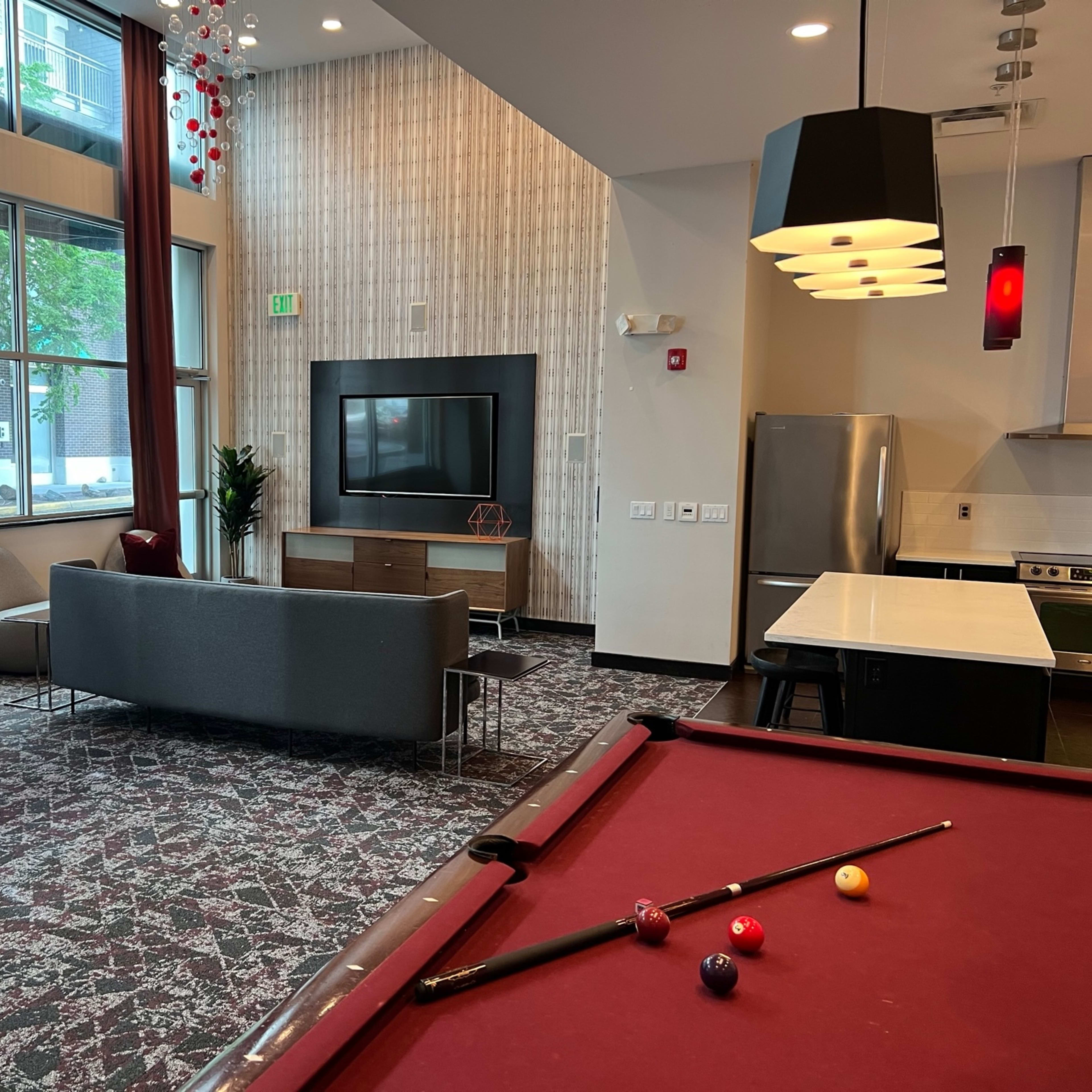 A lounge area features a pool table in the foreground, a modern seating arrangement, and large windows letting in natural light.
