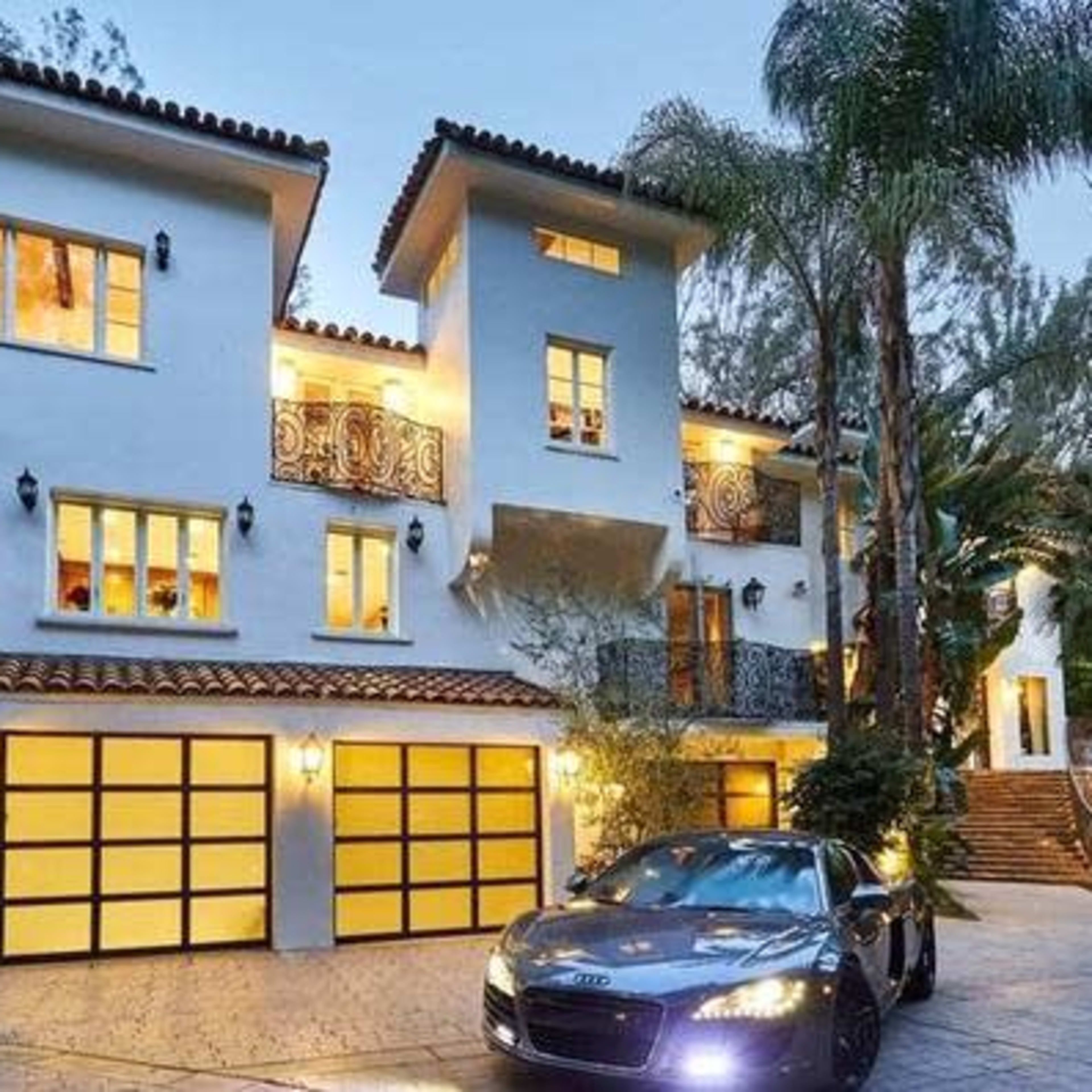 A two-story white stucco house with a tile roof, featuring multiple balconies and a landscaped driveway with a parked black sports car.