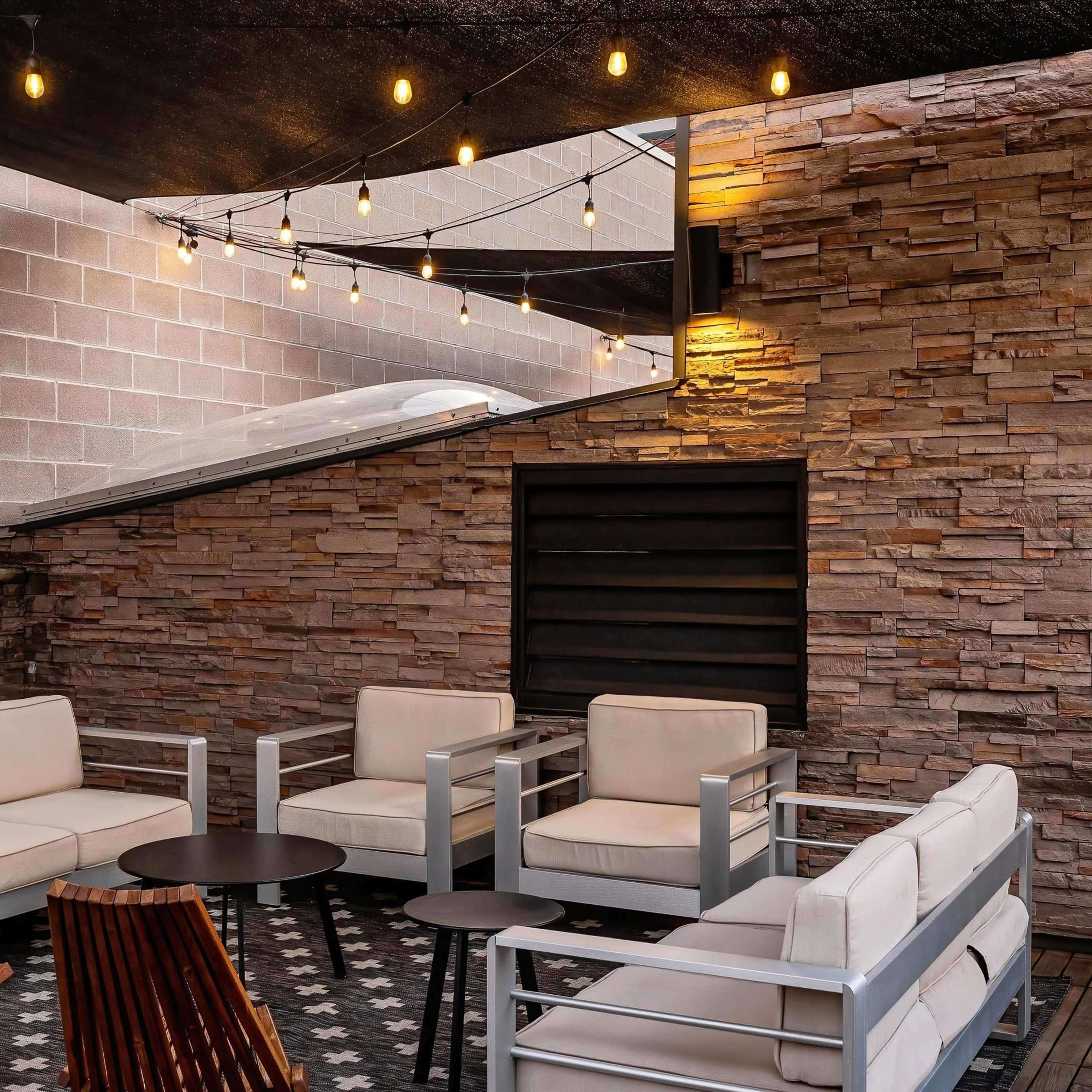 A modern outdoor lounge area featuring beige sofas, a wooden chair, a circular table, and string lights above, set against a stone wall backdrop.