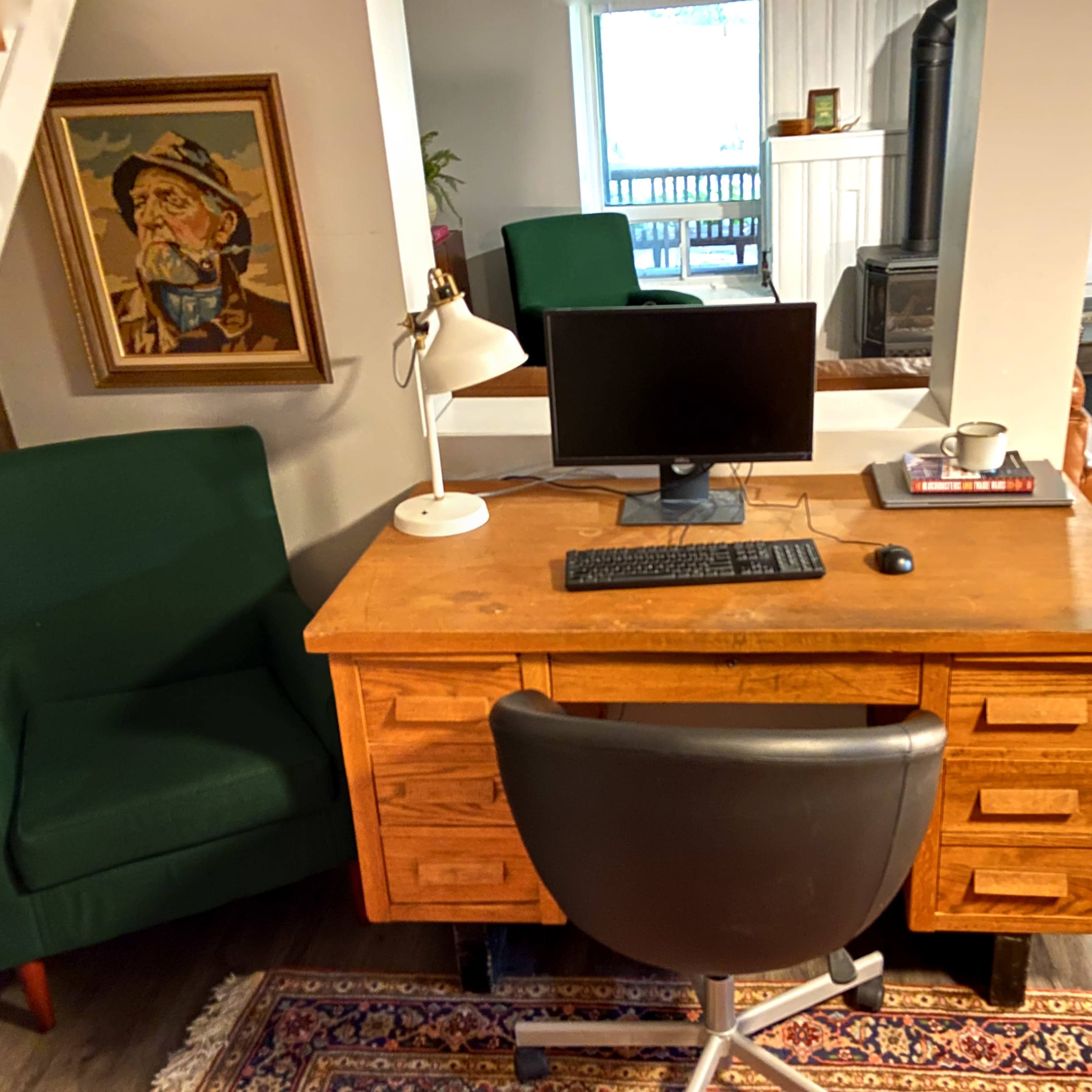 A wooden desk with a computer monitor, keyboard, and a small cup sits in a room featuring a green chair and a doorway leading outside.