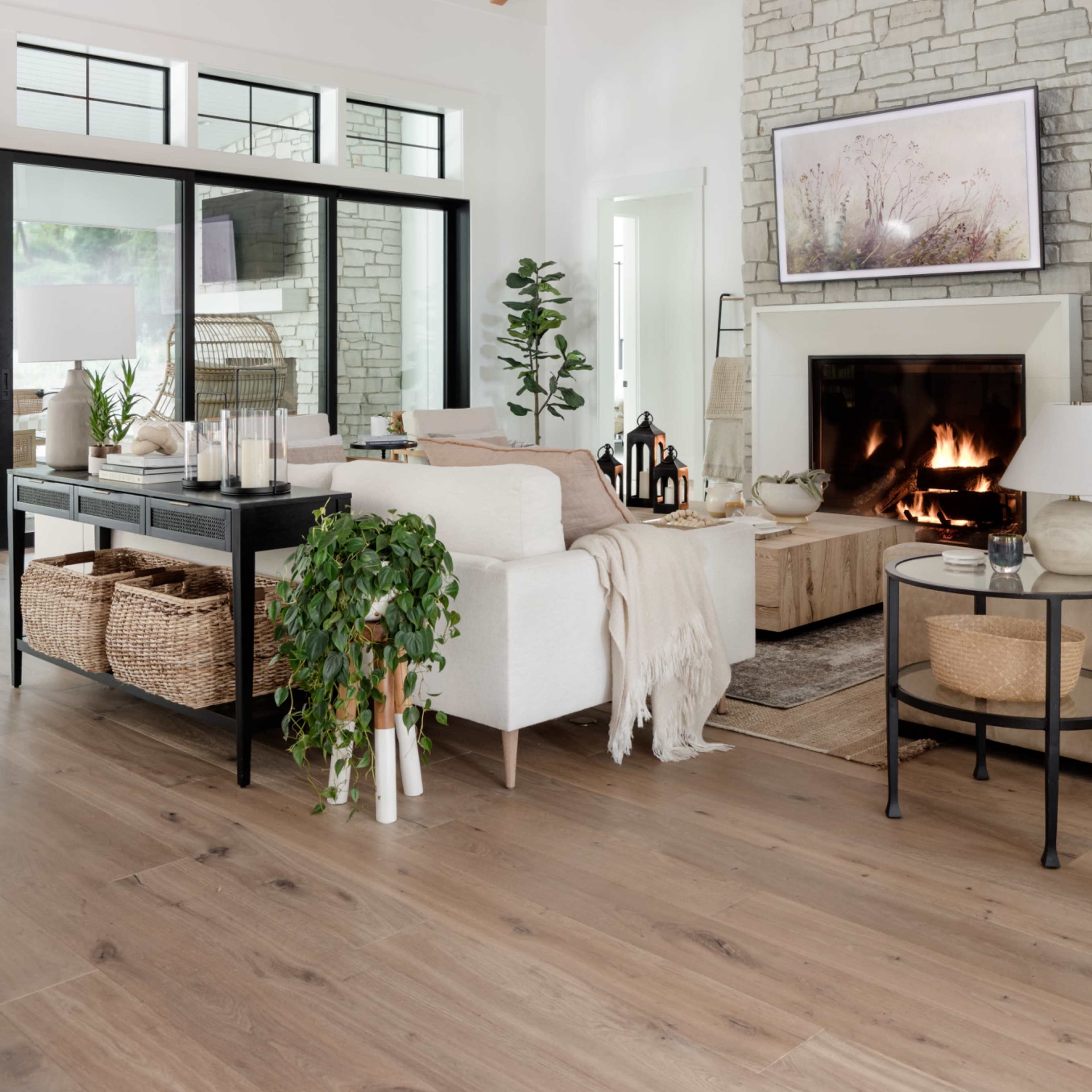 A modern living room features a stone fireplace, high wooden beams, and a white sofa adorned with a throw blanket, alongside decorative elements like plants and woven baskets.
