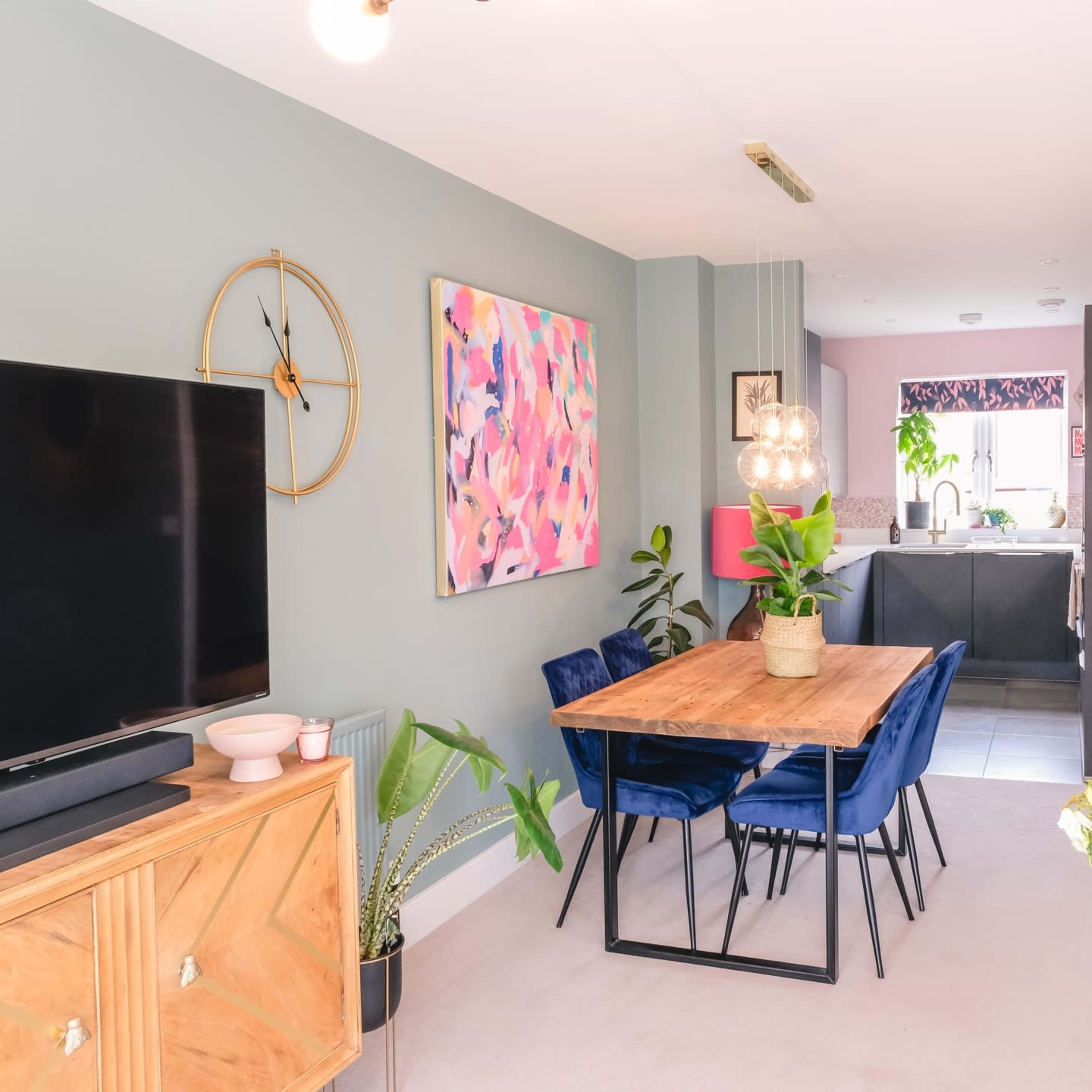 A modern dining area features a wooden table surrounded by blue chairs, with a television mounted on the wall and an open kitchen visible in the background.