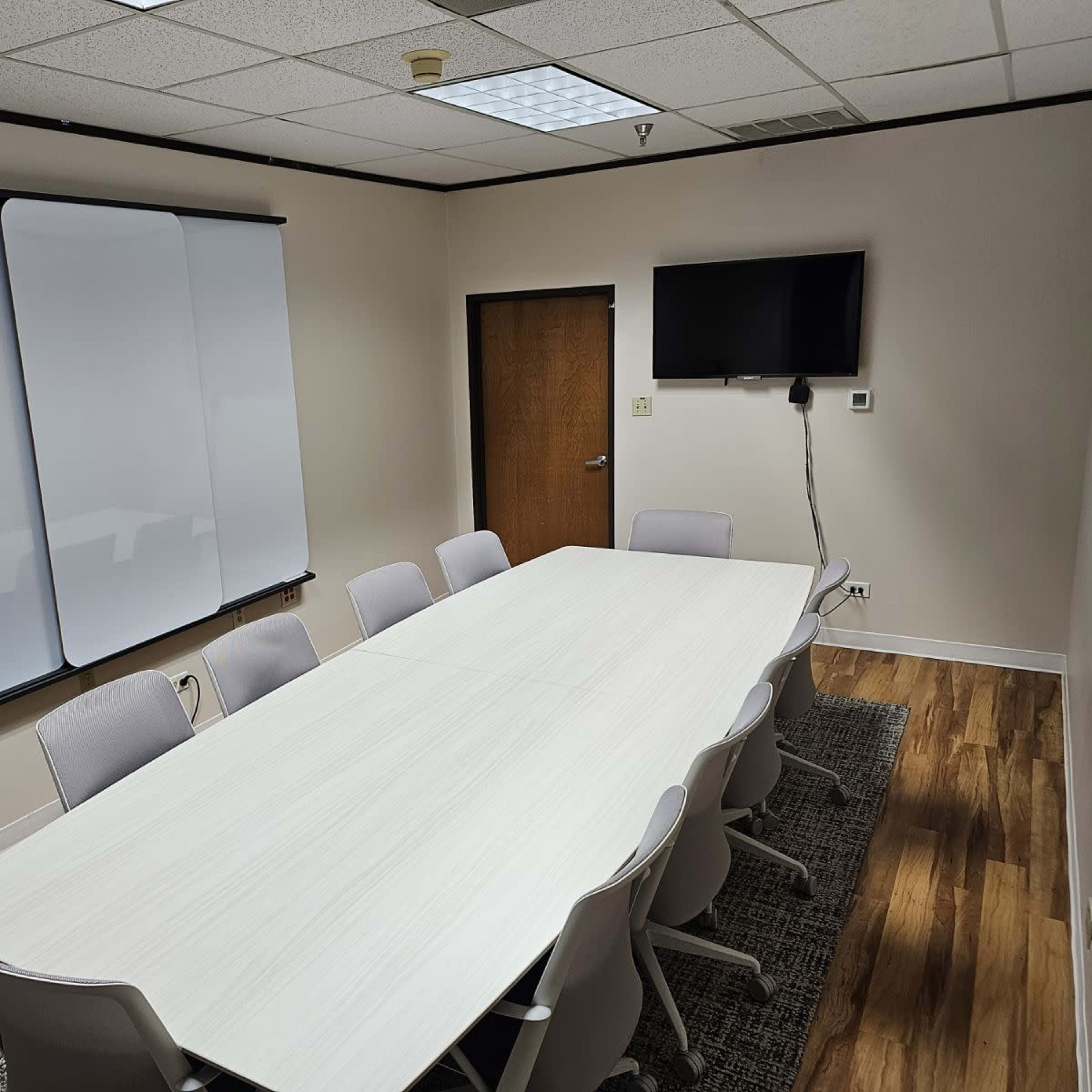 A conference room features a long white table surrounded by gray chairs, a wall-mounted TV, and two whiteboards.