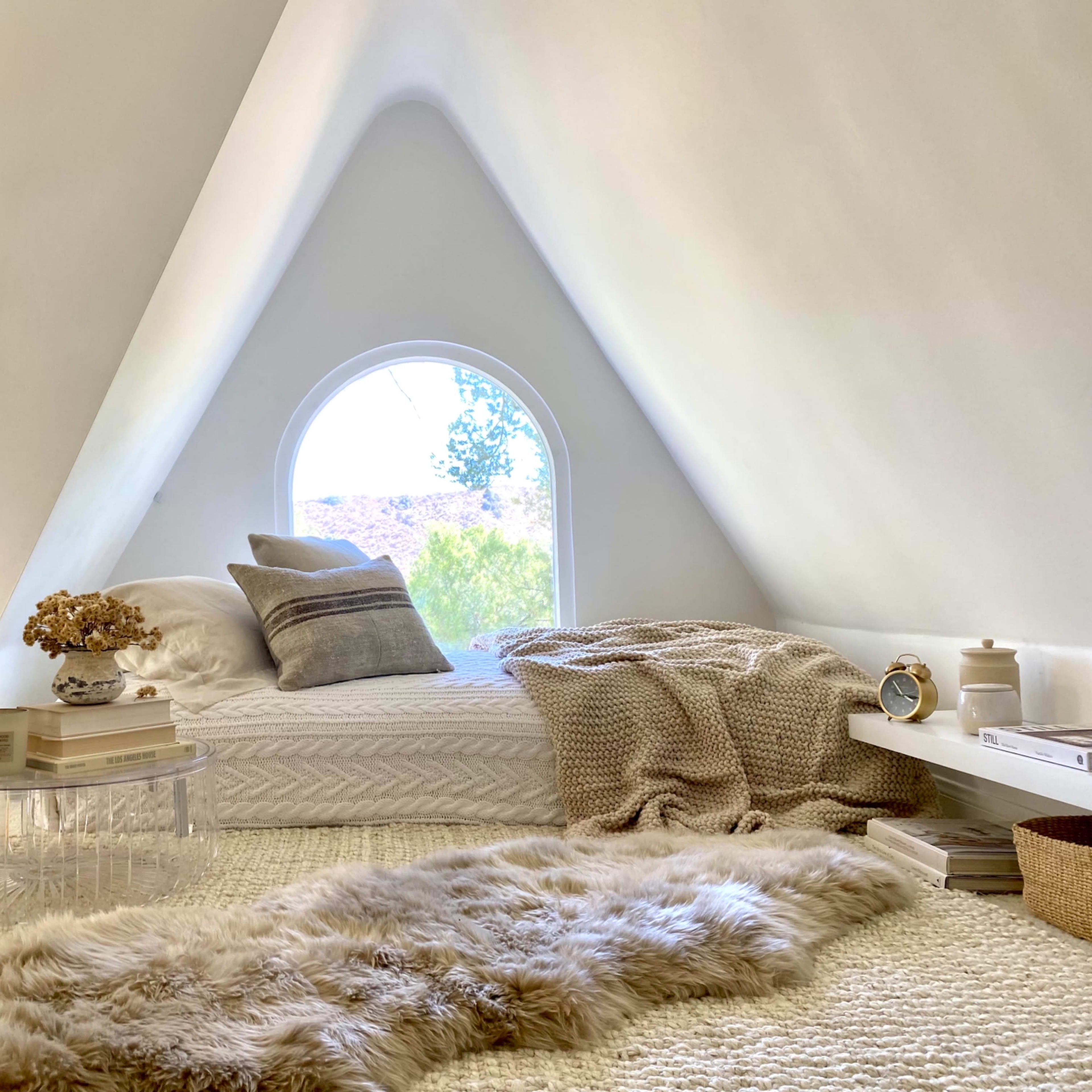 A minimalist bedroom with a sloped, white wall, a large arched window, and a bed adorned with neutral-colored blankets and decorative items.